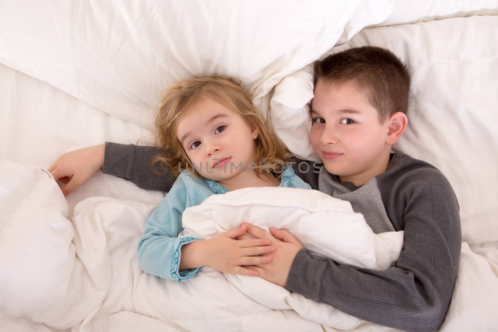 Affectionate little brother and sister lying in bed cuddling together under the duvet looking up sleepily at the camera