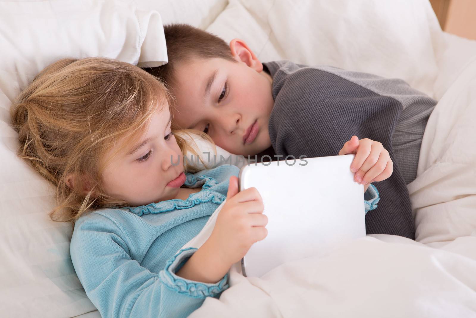 Cute little girl and boy reading a bedtime story as they lie together in bed looking at a tablet computer as they prepare to go to sleep with the little sister holding the notebook