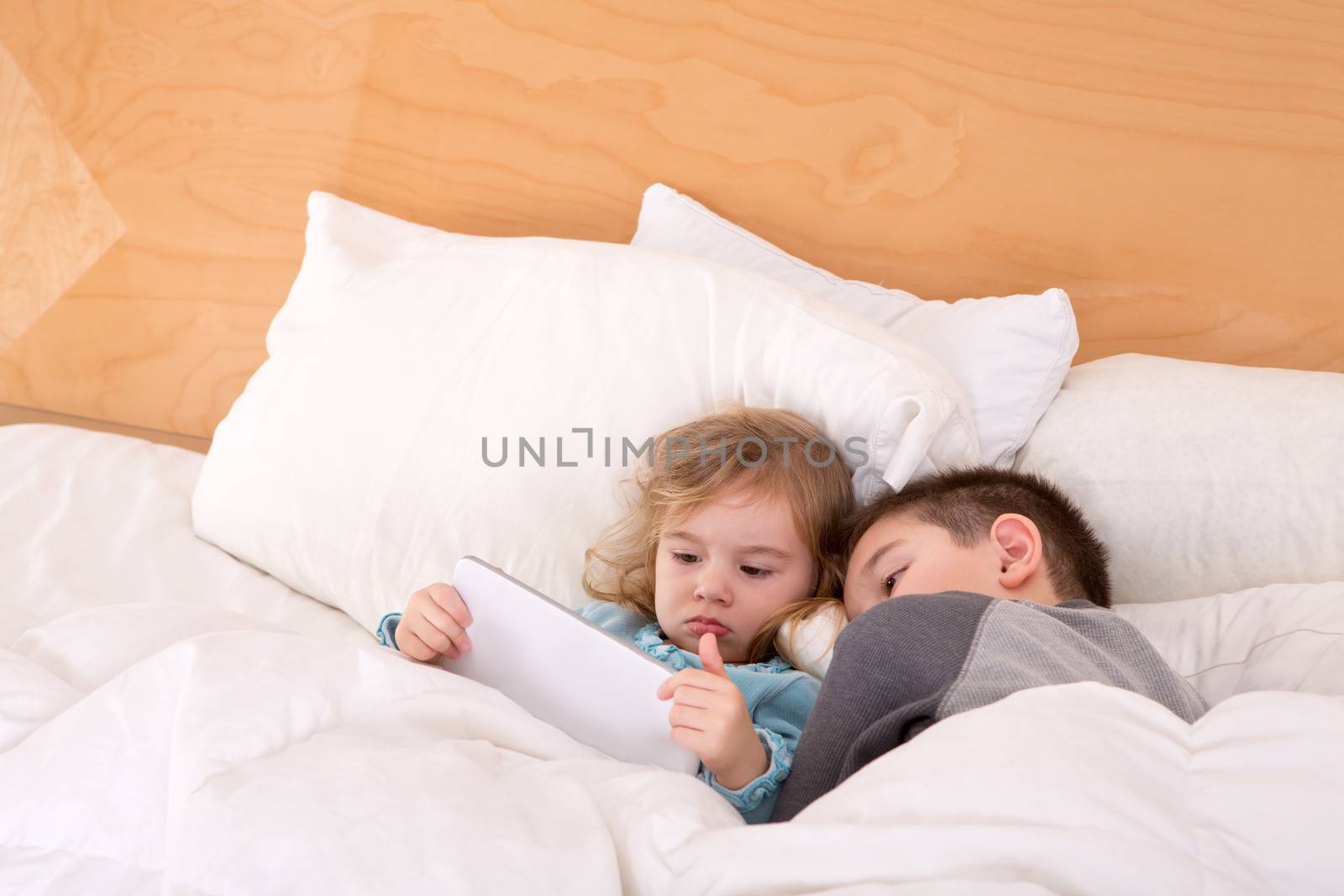 Tired little brother and sister snuggling up together in a warm comfortable bed as they read a bedtime story on a tablet computer before falling asleep