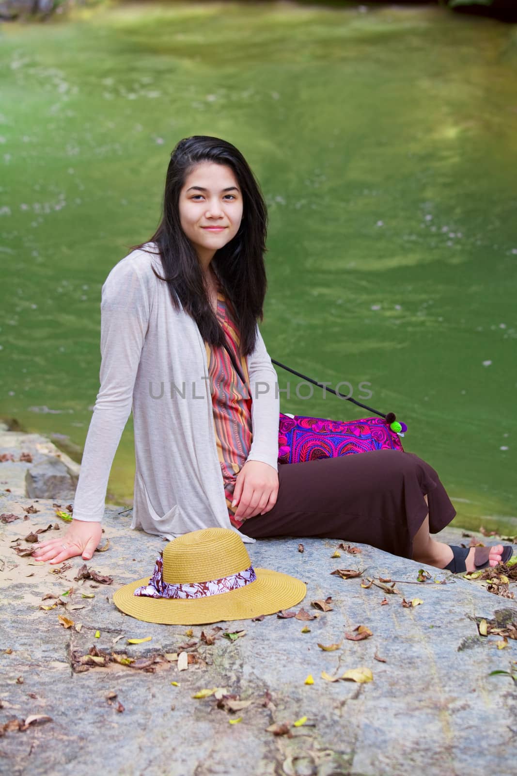 Biracial teen girl sitting next to river, smiling