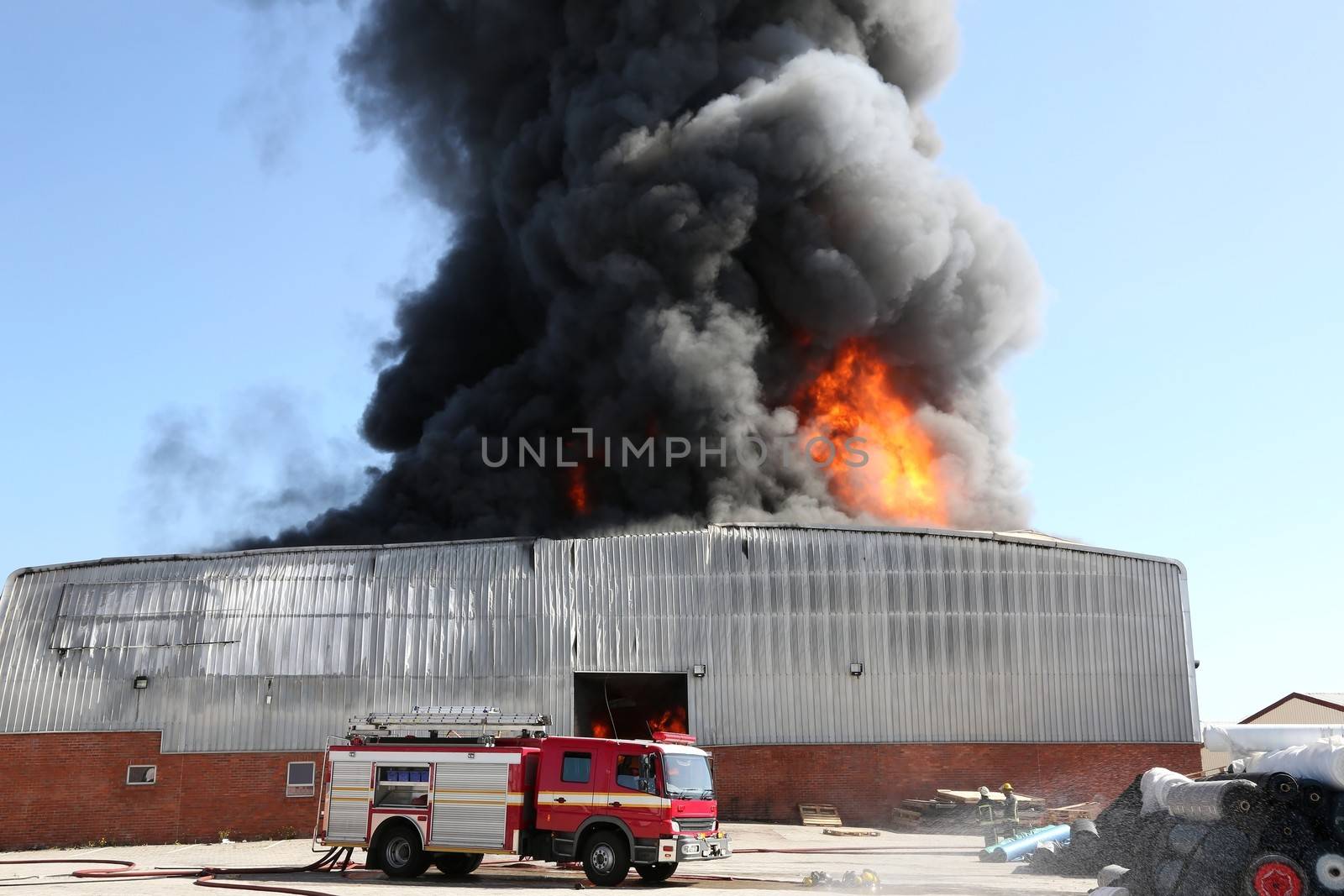 Warehouse building burning with intense flames and firemen attending
