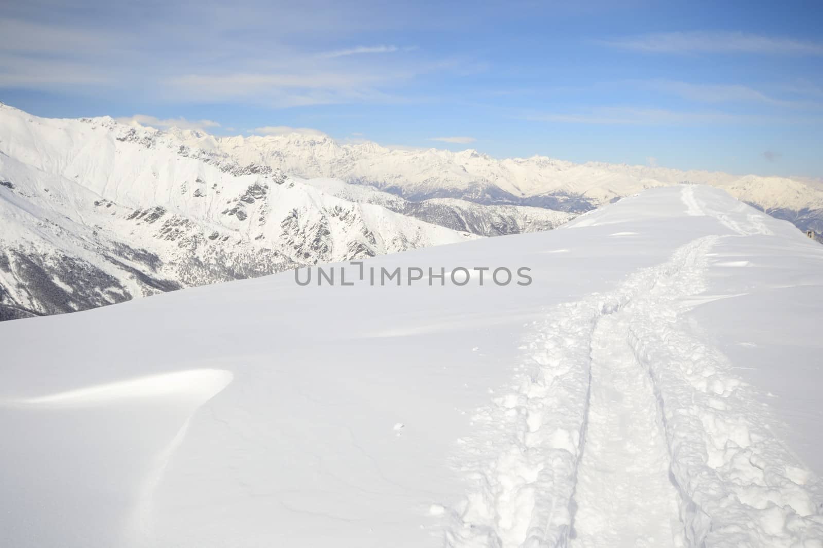 Alpinist hiking uphill by ski touring on the mountain ridge in powder snow with deep track in the foreground and scenic high mountain view in the background