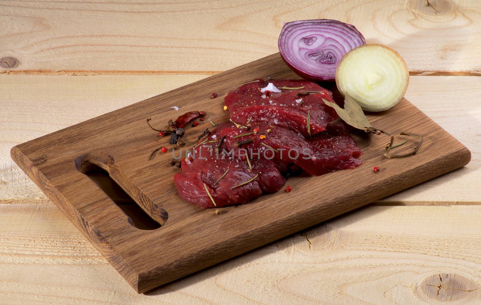 Arrangement of Raw Sliced Beef with Halves of White and Red Onion, Bay Leafs and Spices on Cutting Board isolated on Wooden background