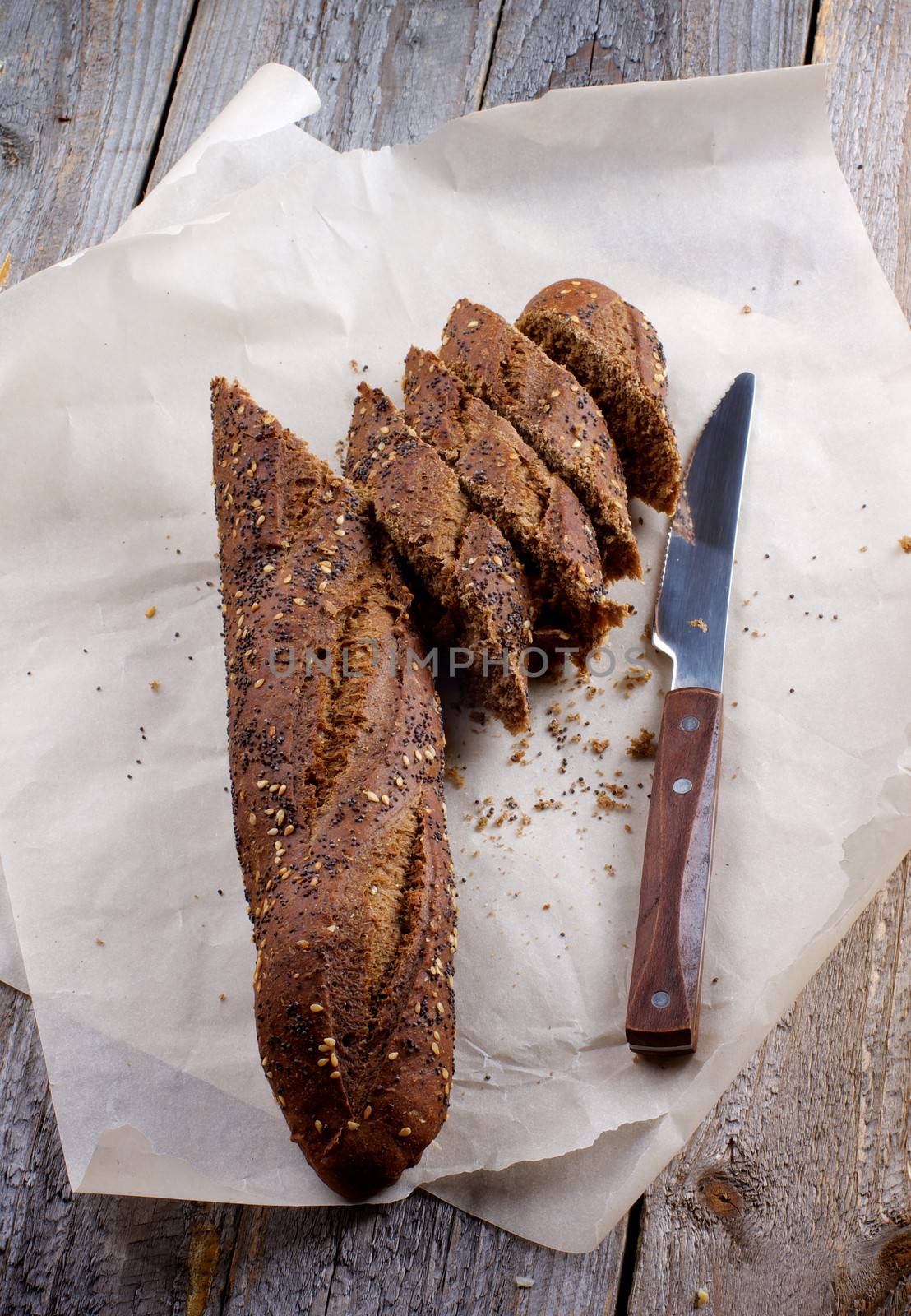 Half of Sesame and Poppy Seed Brown Bread and Slices with Table Knife on Parchment Paper closeup on Wooden background 