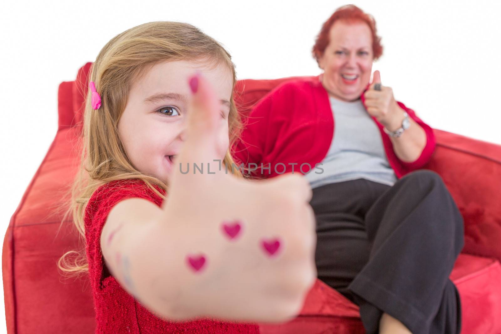 Cute little girl with red hearts painted on her hand giving a thumbs up gesture of approval with her grandmother who is seated on a red sofa behind her