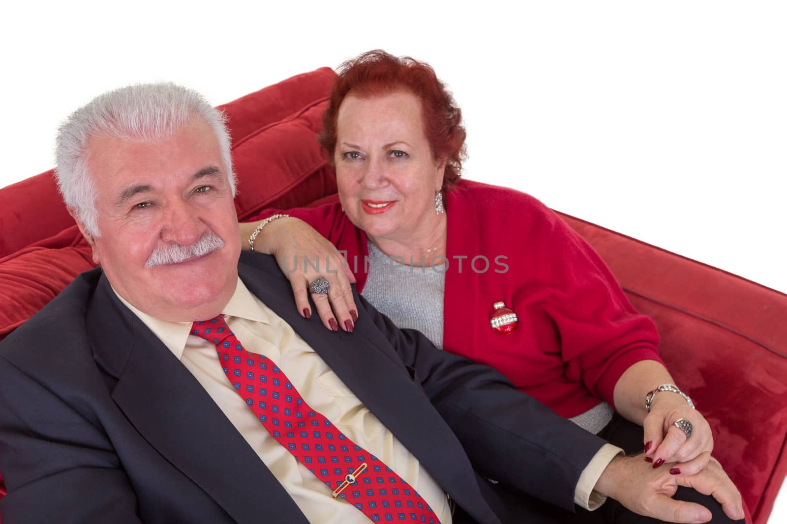 High angle view of an attractive stylish senior couple seated together on a red couch looking up at the camera with friendly smiles on a white background