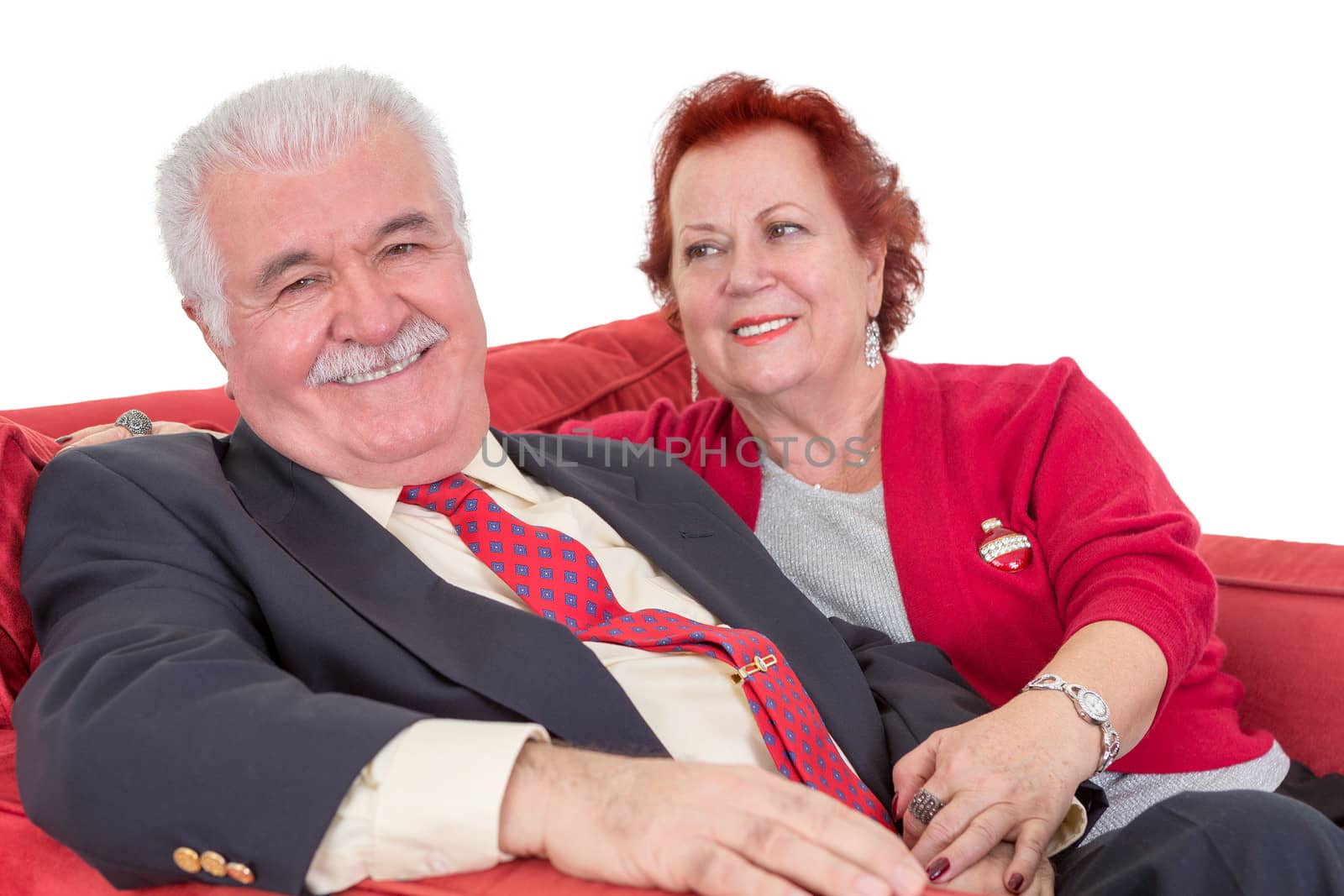 Devoted senior couple seated on a red sofa holding hands with the wife looking lovingly at her husband as he gives the camera a beaming smile