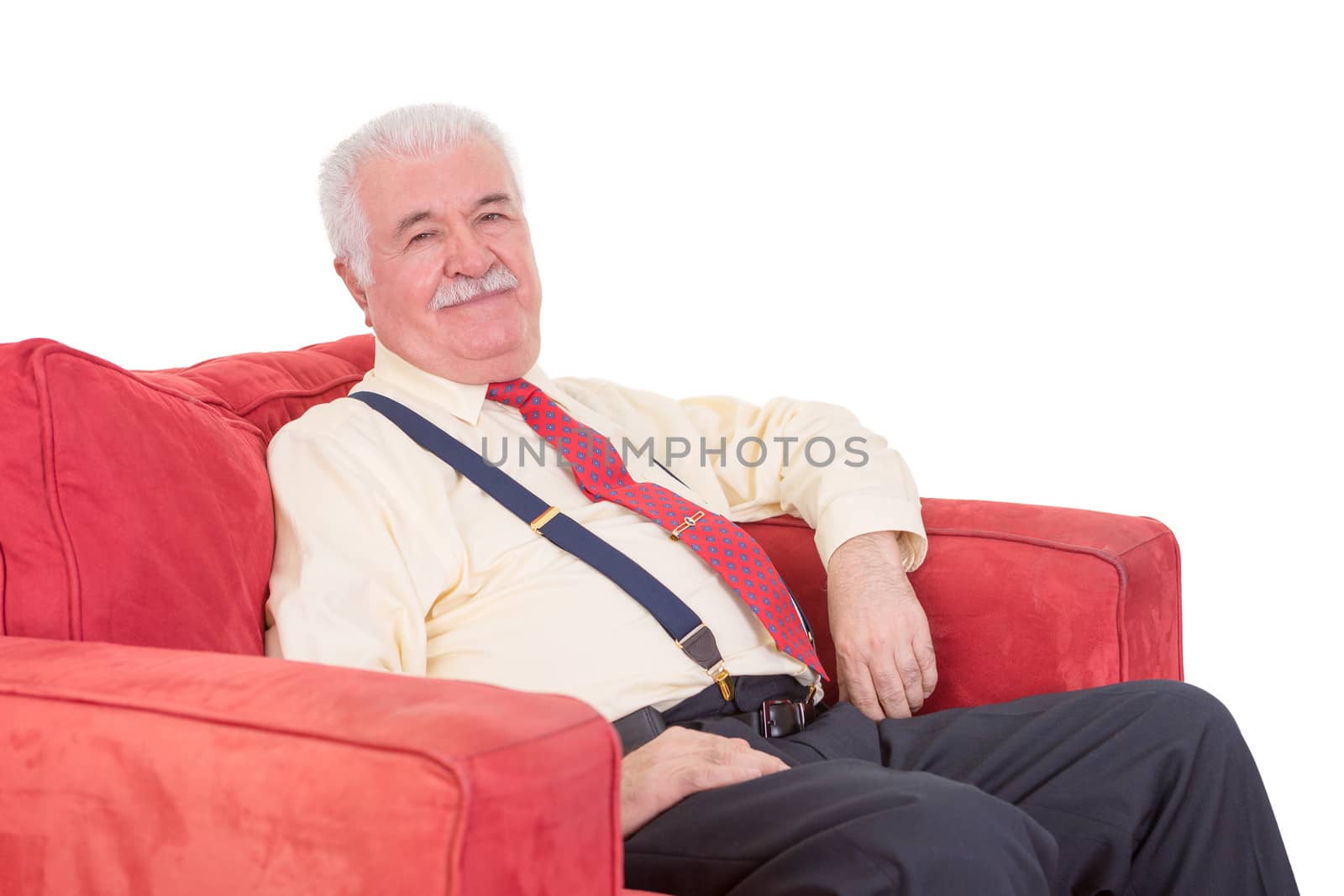 Senior grey-haired gentleman with a moustache wearing braces relaxing in a comfortable red armchair on a white background