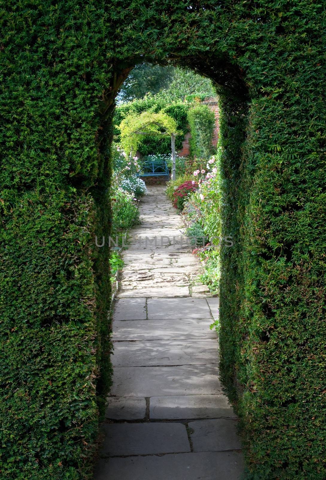 Pretty flagstone path through clipped yew archway towards a bench with flowers, Cotswolds, England.