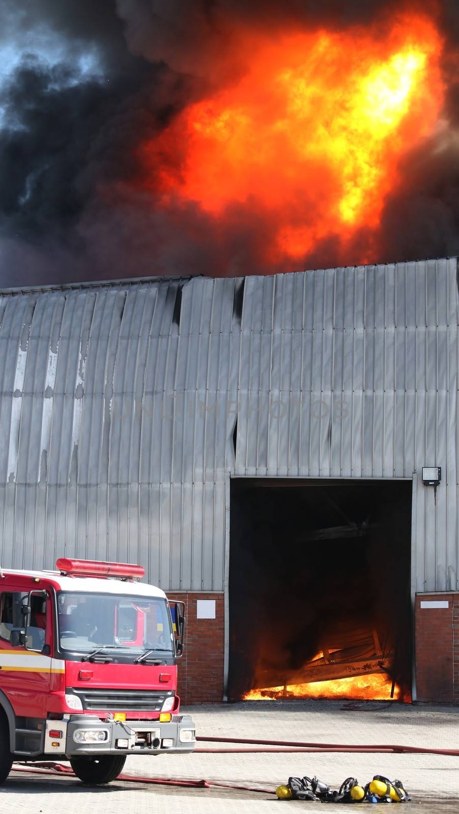 Warehouse building burning with intense flames and firemen attending