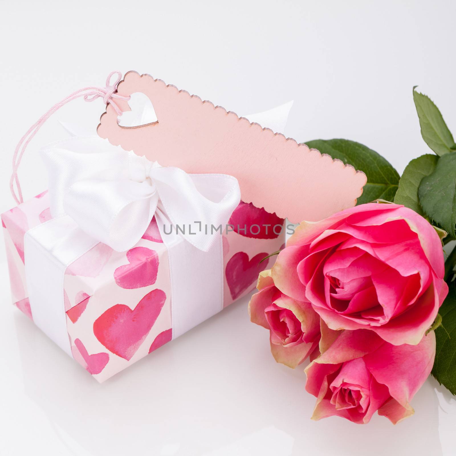 Close-up of a gift box wrapped in decorative paper with pattern with heart shapes, with an empty tag, next to three roses, symbol of love and appreciation