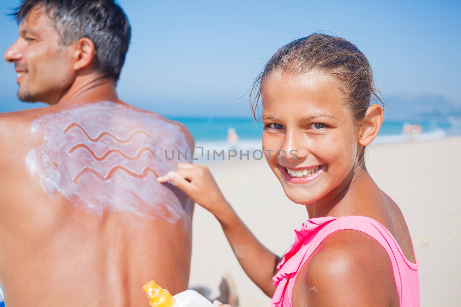 Adorable girl at tropical beach applying sunblock cream on a father's back.