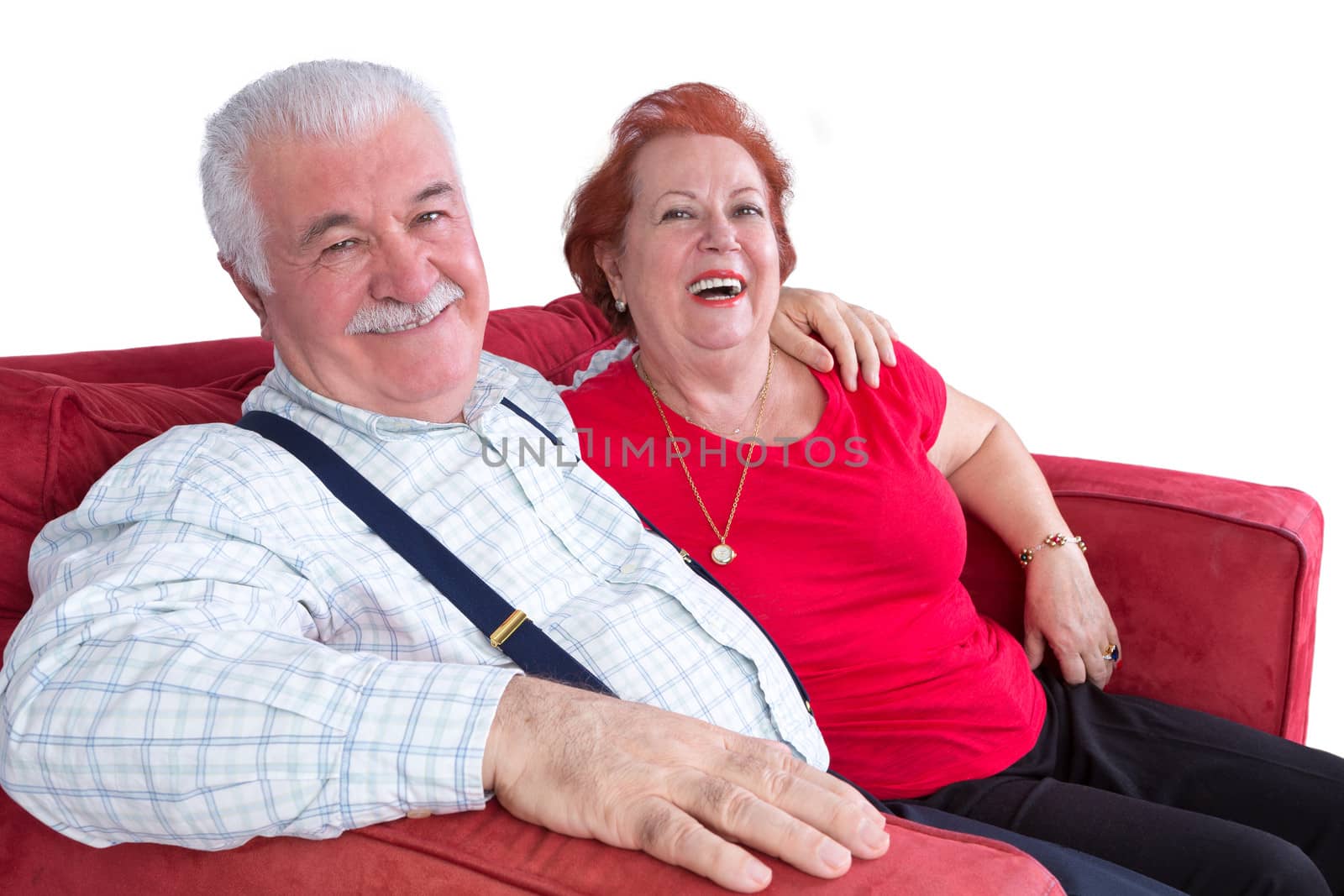 Joyful relaxed elderly couple sitting arm in arm on a red sofa laughing at the camera over white
