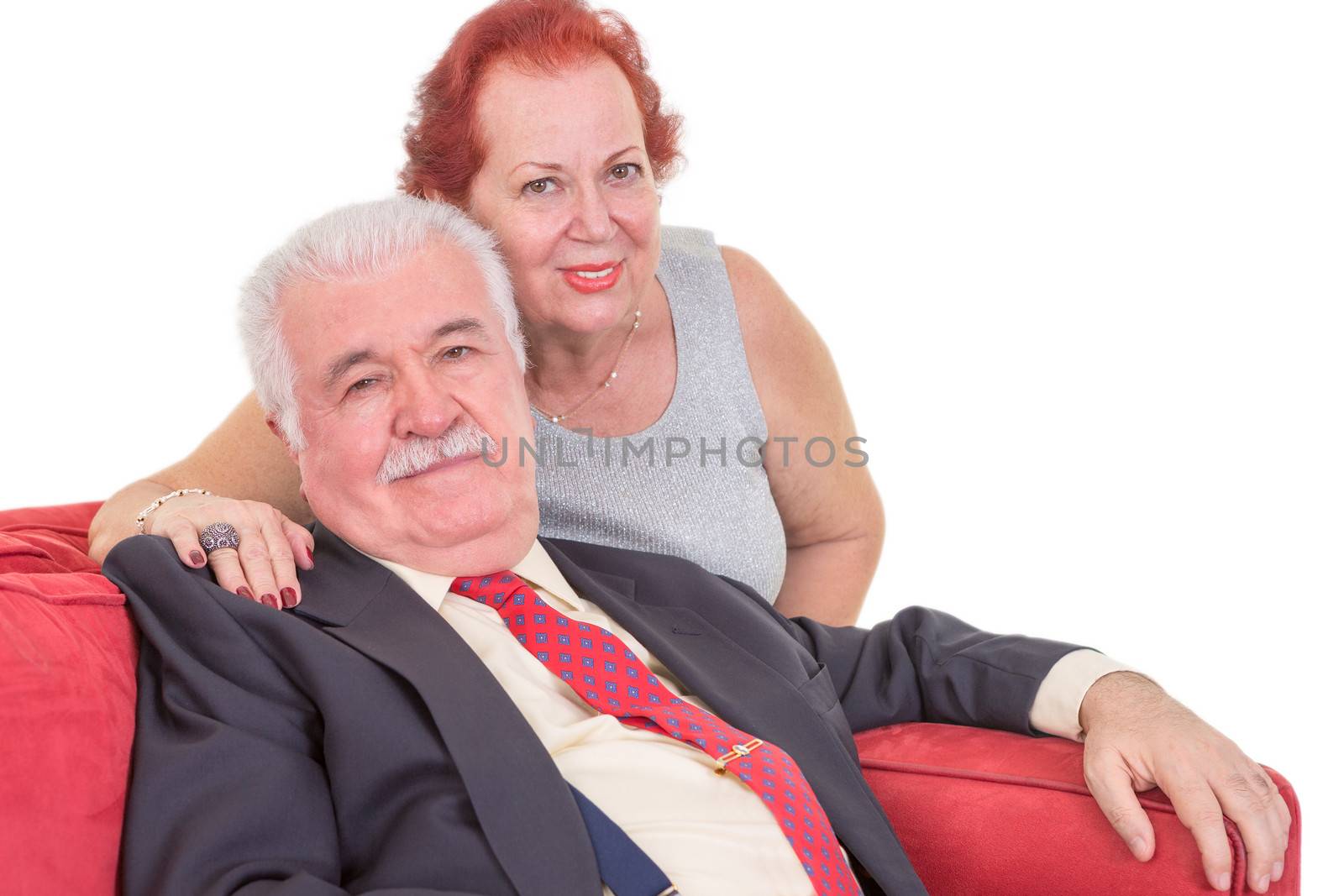 Loving elderly couple relaxing together on a red couch with the wife sitting behind her husband with her arm around his shoulders looking at the camera