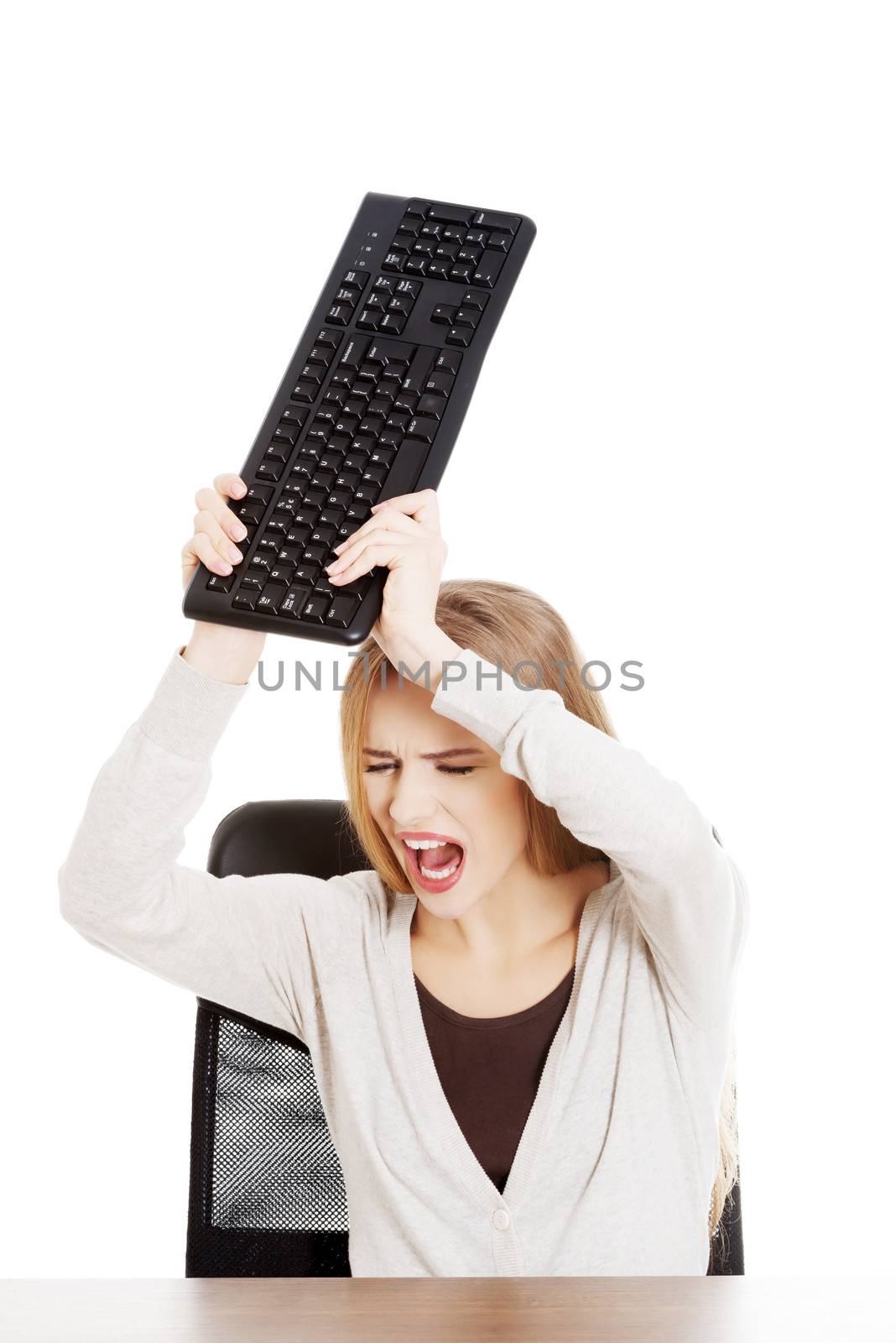 Beautiful casual woman behind the desk trying to broke the keyboard. Isolated on white.