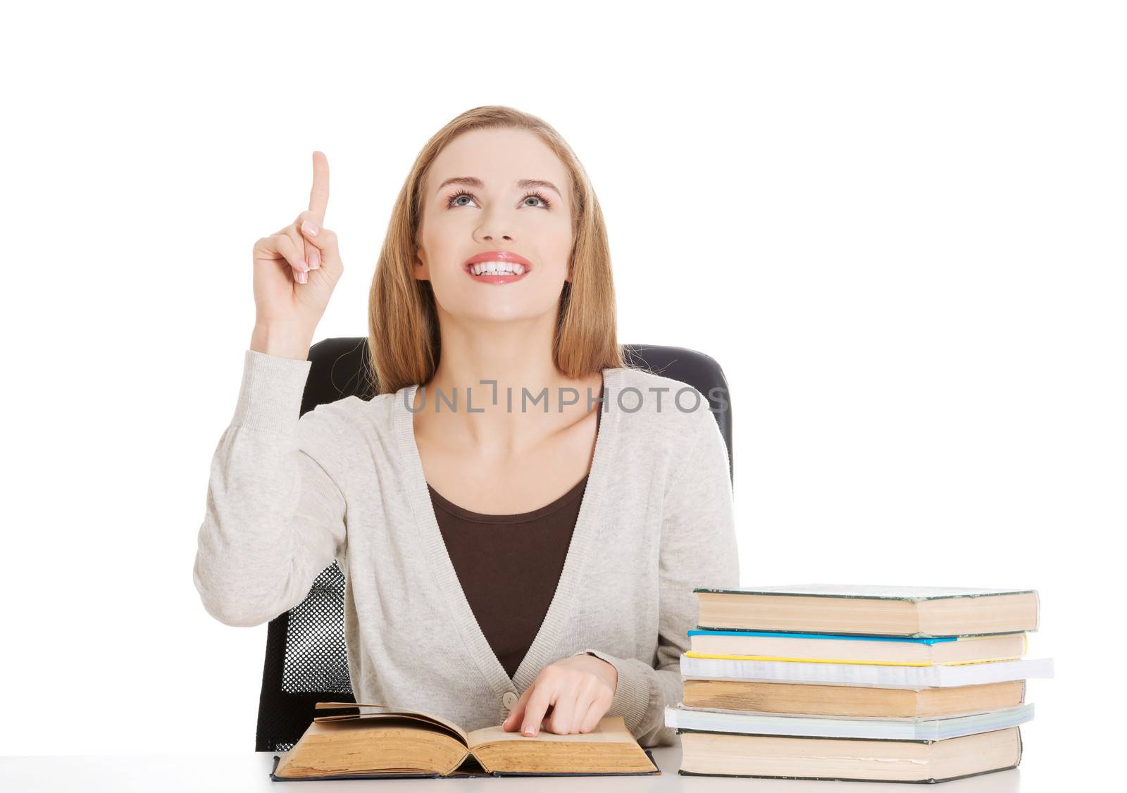 Beautiful casual student woman sitting by stack of books and pointing up, experessing happiness and satisfaction. Isolated on white.