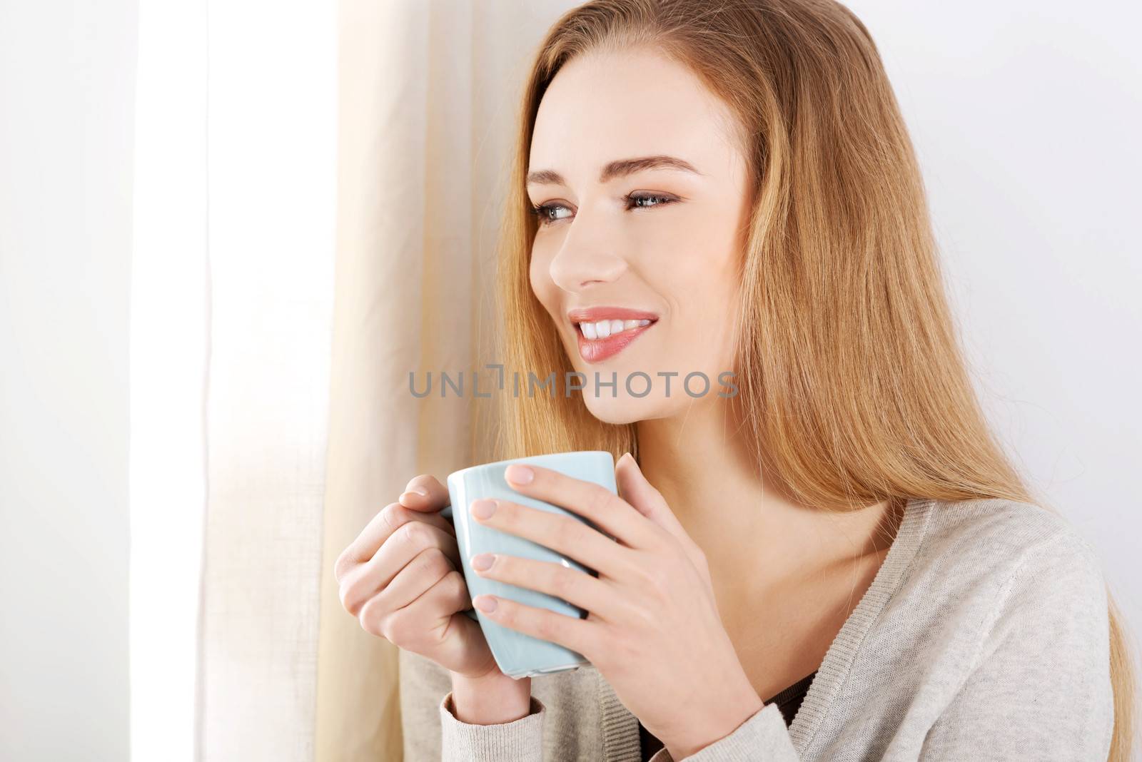 Young beautiful woman holding a cup and looking through window. Indoor background.