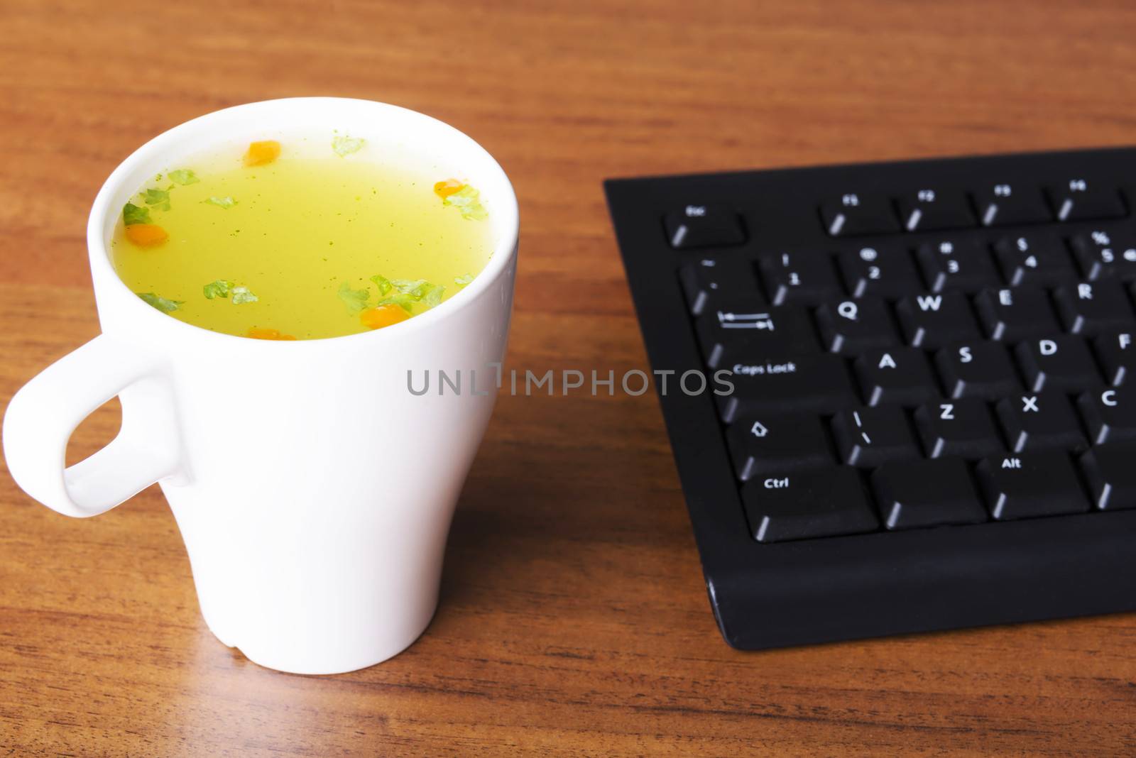 Hot vegetable soup in a white cup next to keyboard standing on wooden table.