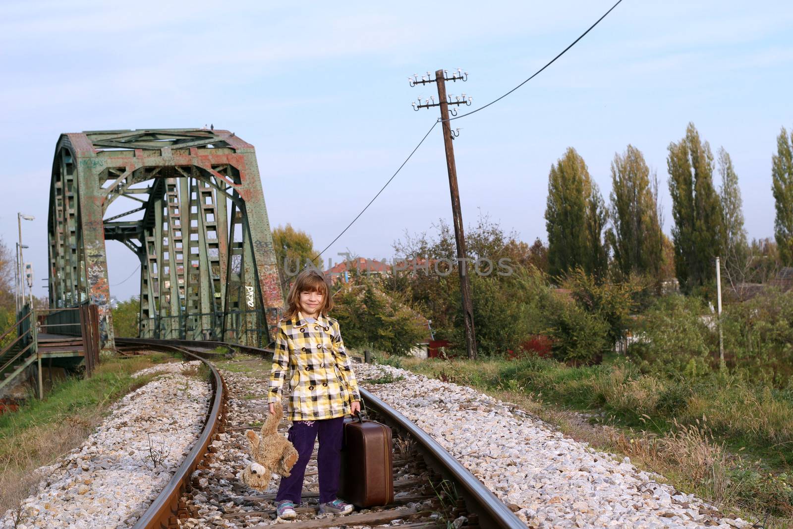 little girl with suitcase and teddy bear standing on railroad by goce