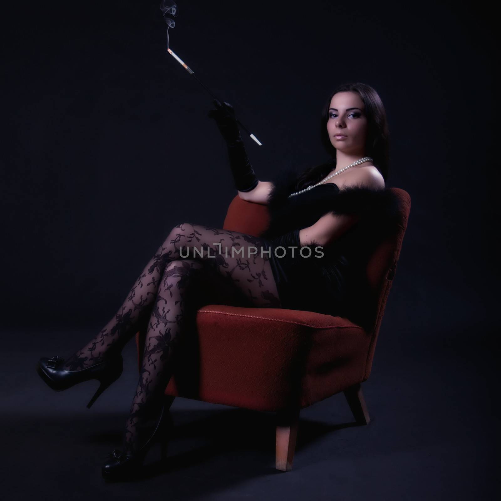 Young woman in retro dress with cigarette holder sitting on an old chair on dark background