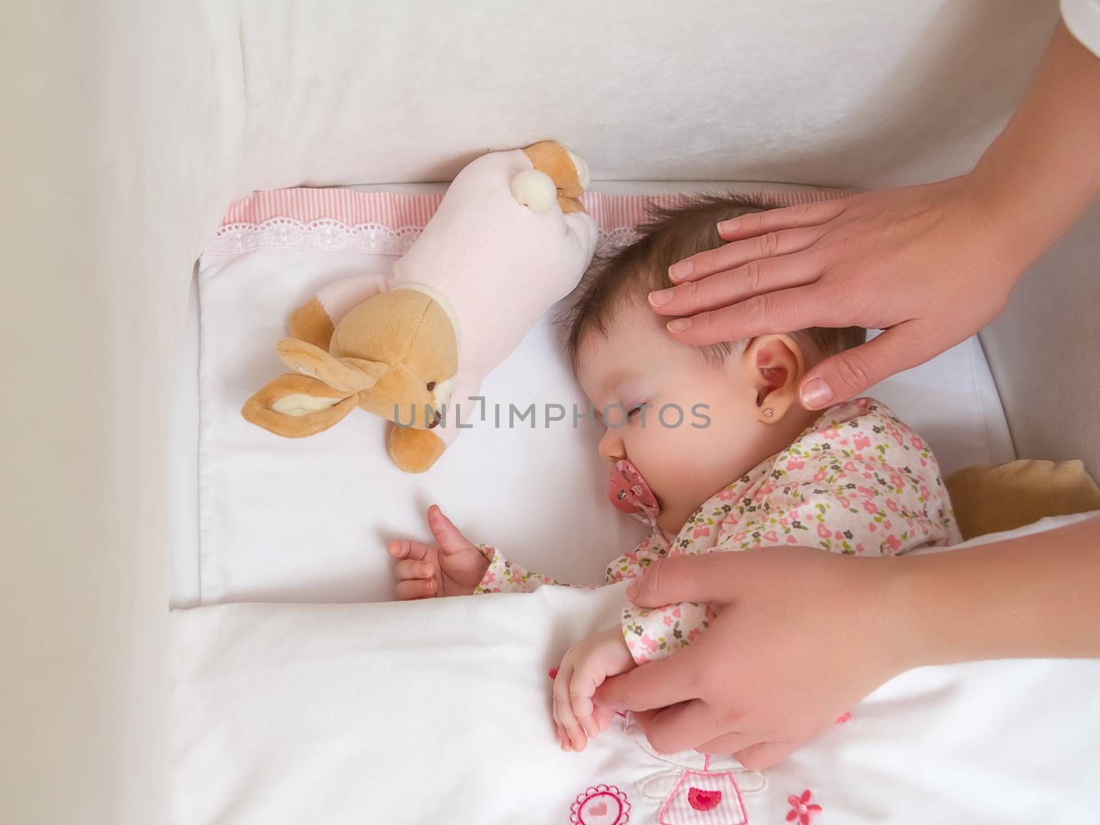 Hands of mother caressing her cute baby girl sleeping in a cot with pacifier and stuffed toy