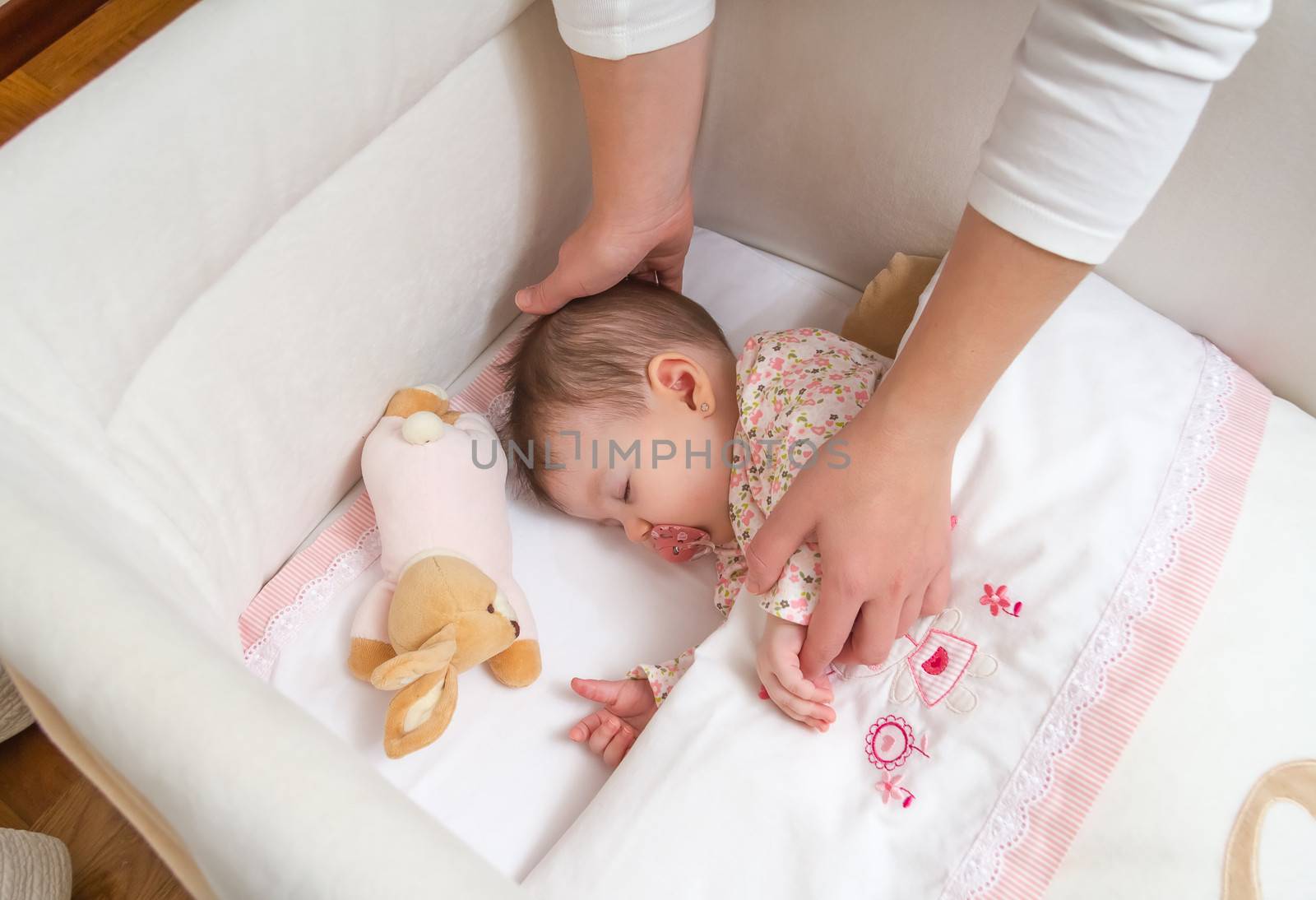 Hands of mother caressing her cute baby girl sleeping in a cot with pacifier and stuffed toy