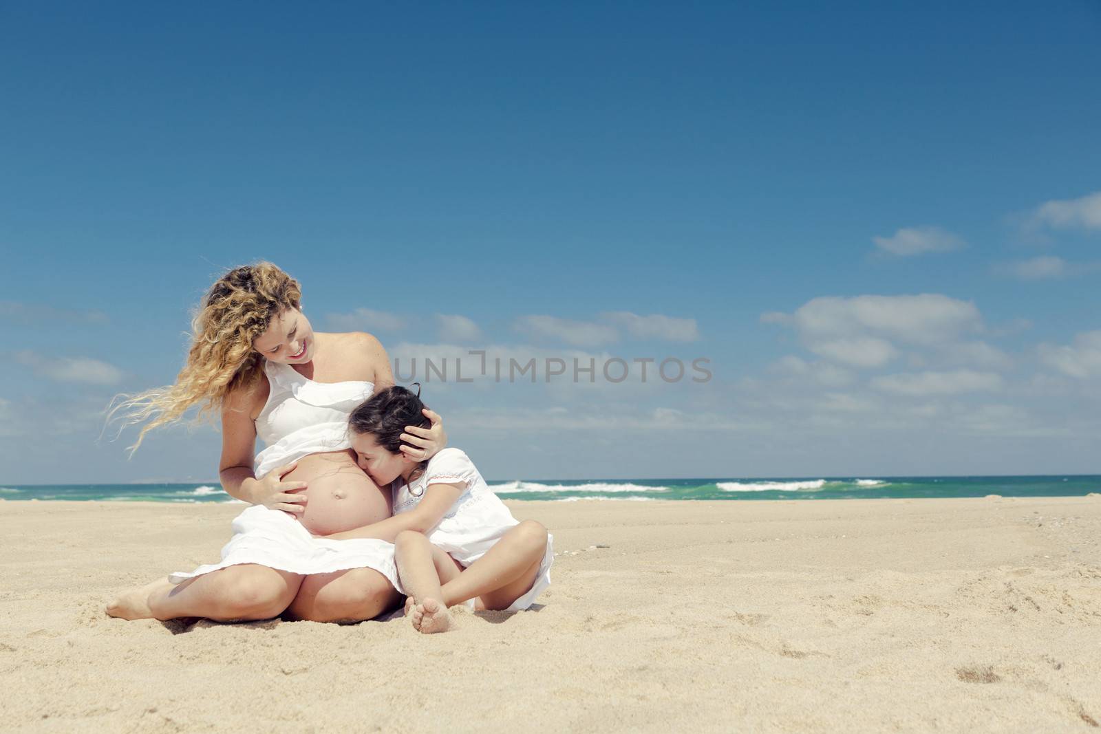 Beautiful pregnant woman and her little daughter kissing mom's belly