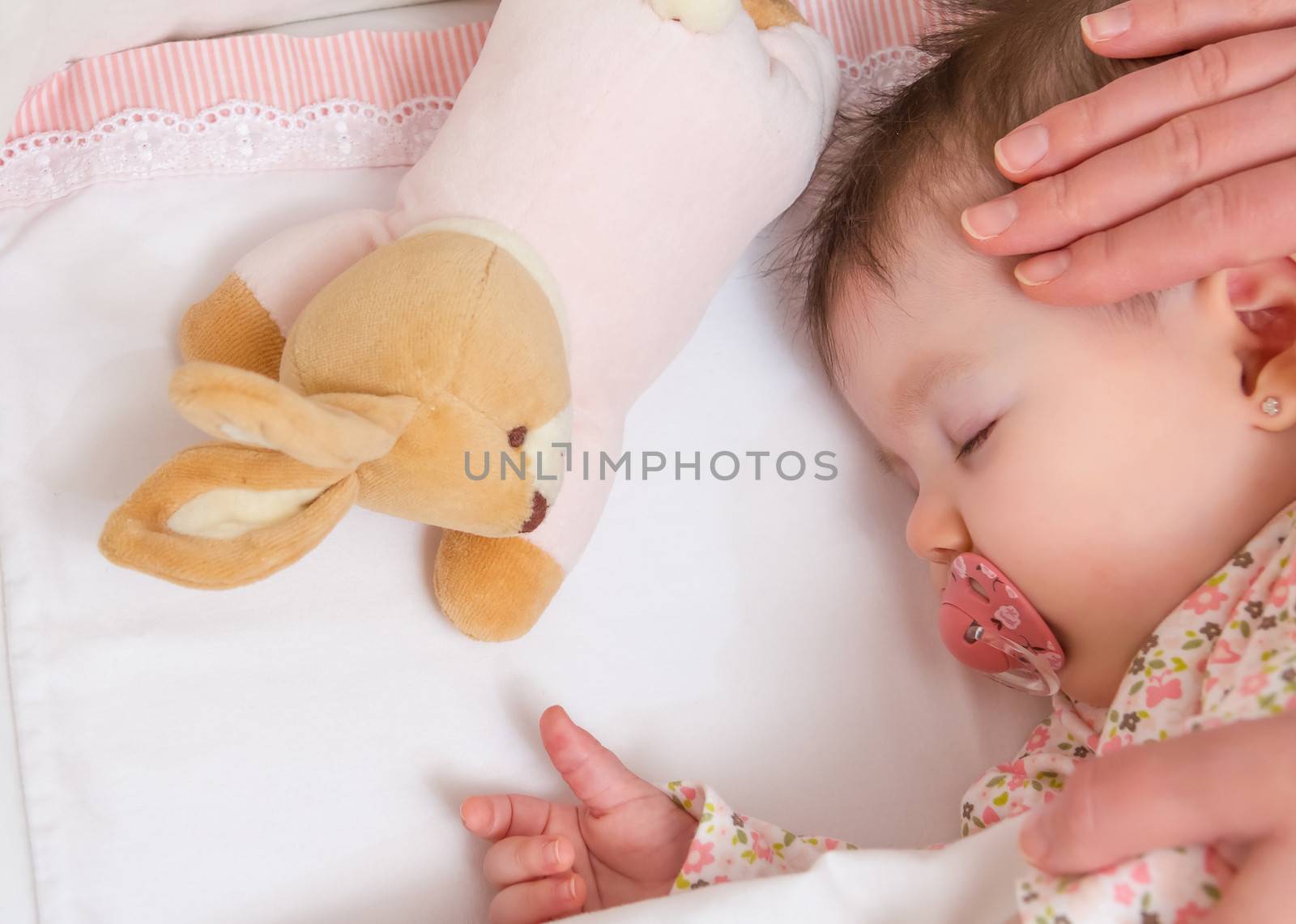 Hands of mother caressing her cute baby girl sleeping in a cot with pacifier and stuffed toy