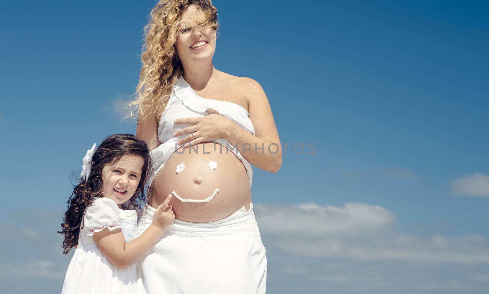 Beautiful pregnant woman in the beach with her little daugther making a smile on mom's belly with sunscreen
