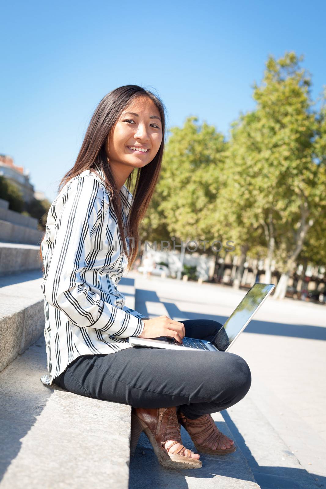 Happy asian girl using a laptop sitting on stairs in a park 