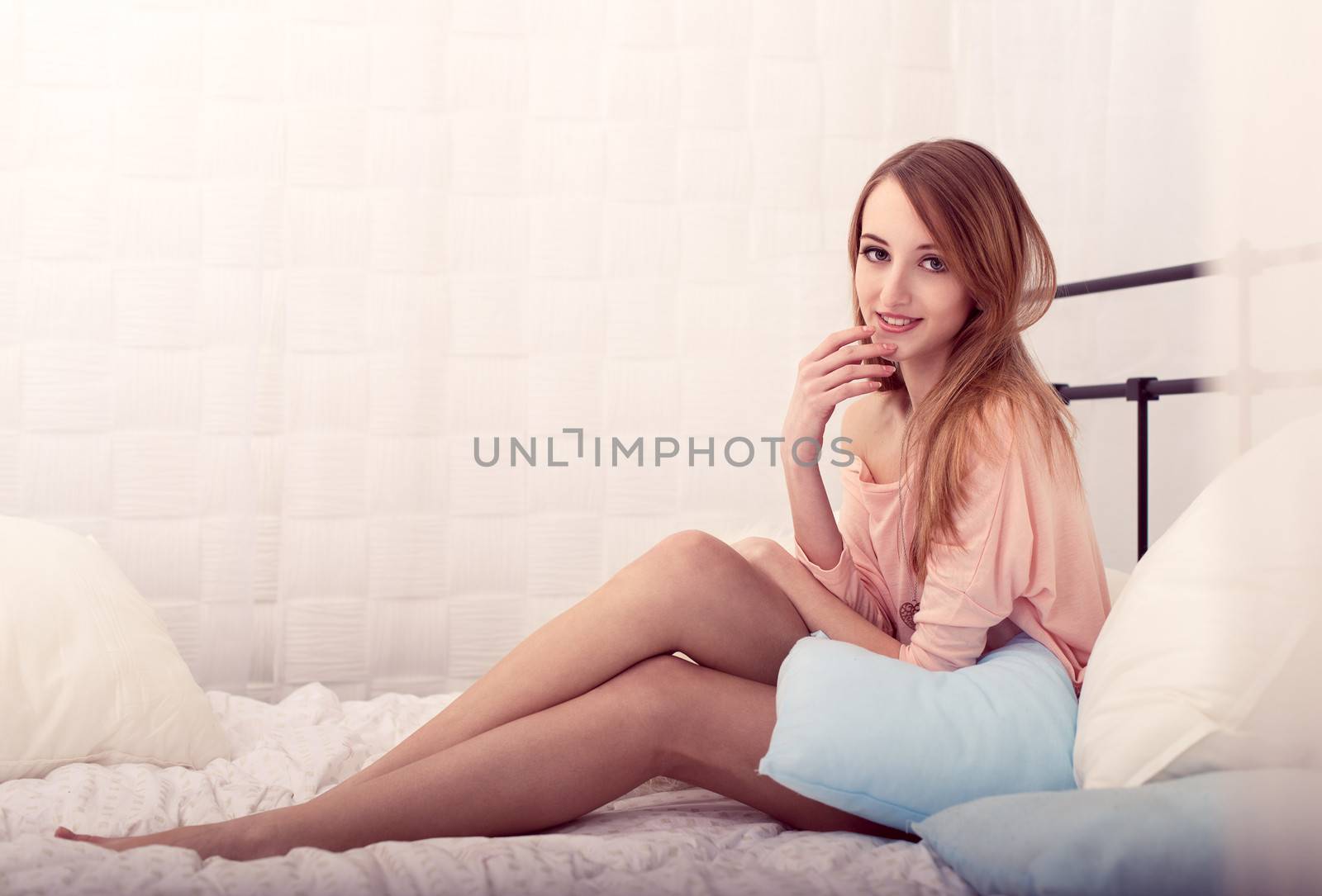 The beautiful young girl in a pink jacket smiles, holding a hand at a mouth, sitting on a bed