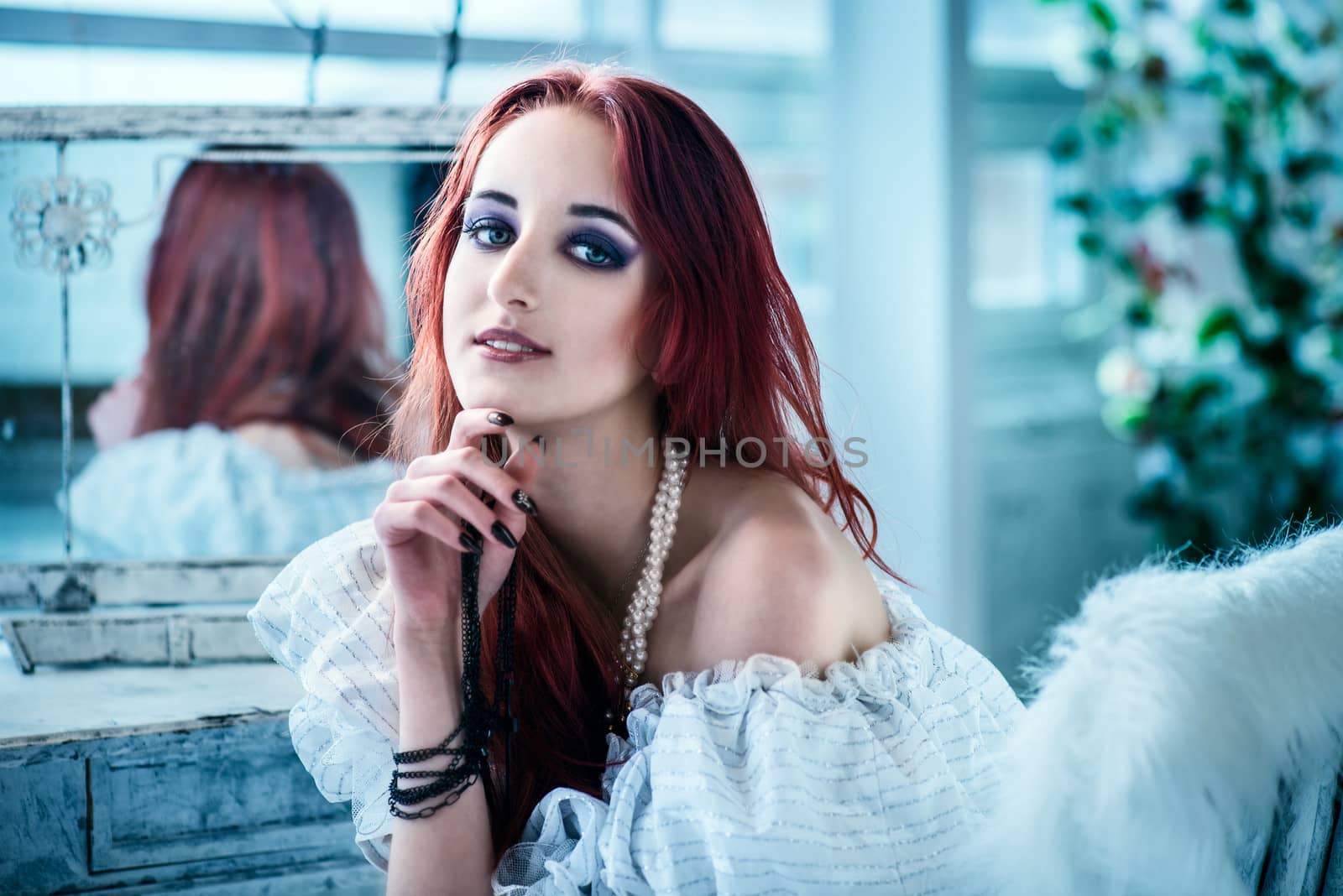 the beautiful girl with red hair in a white dress sits a back to a mirror, having added a hand under a chin
