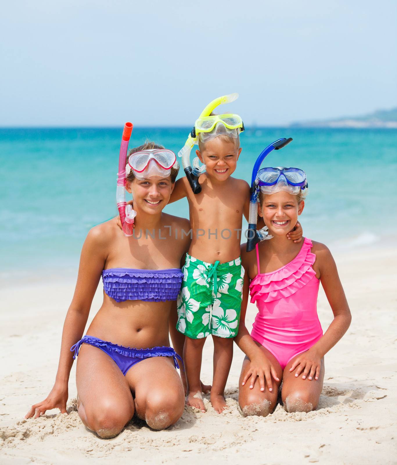 Three happy children on beach with colorful face masks and snorkels, sea in background.