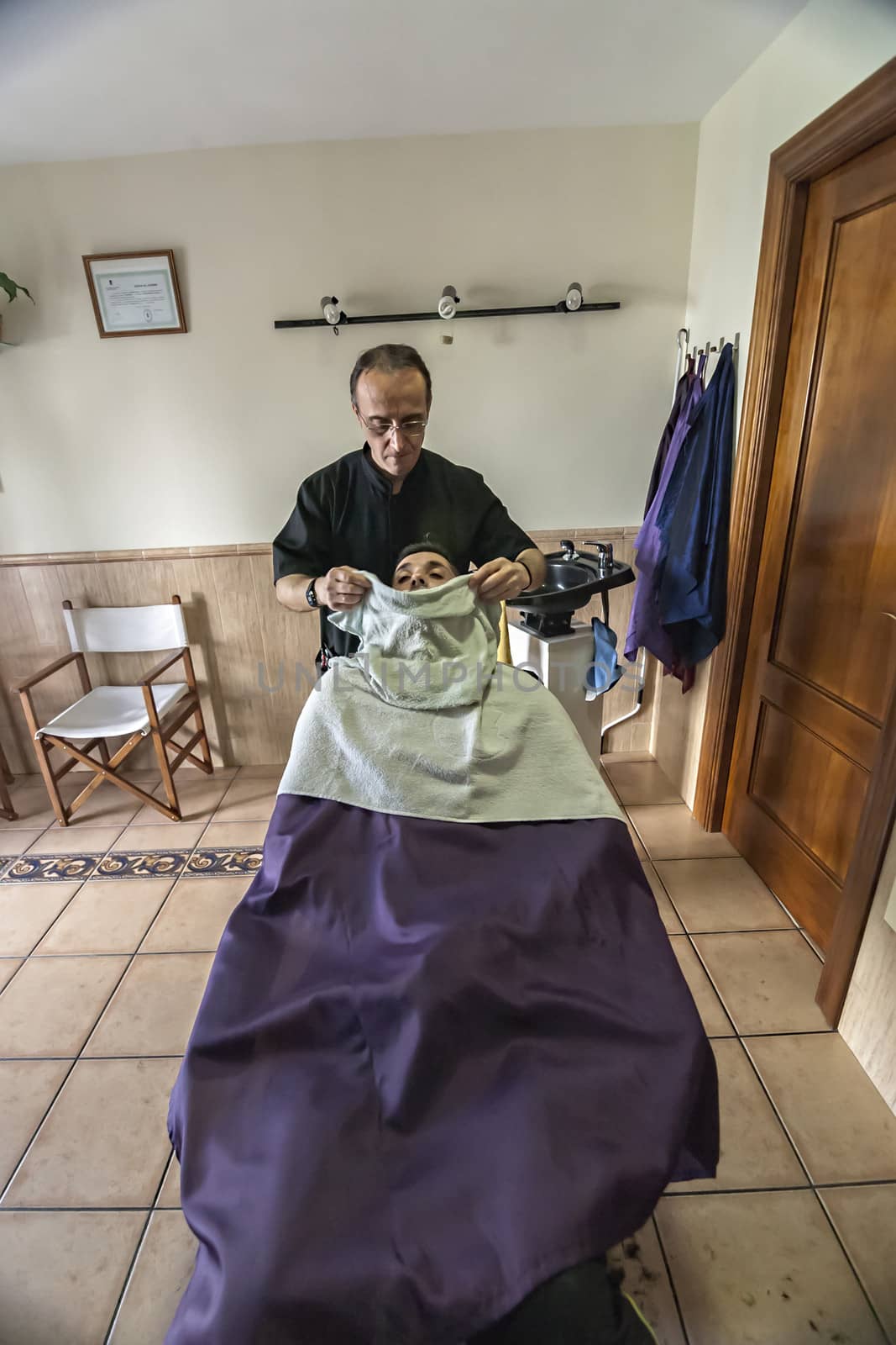 barber applies a hot water cloth in the face of the client to relax the skin in a barber's shop, Sabiote, Jaen province, Andalucia, Spain