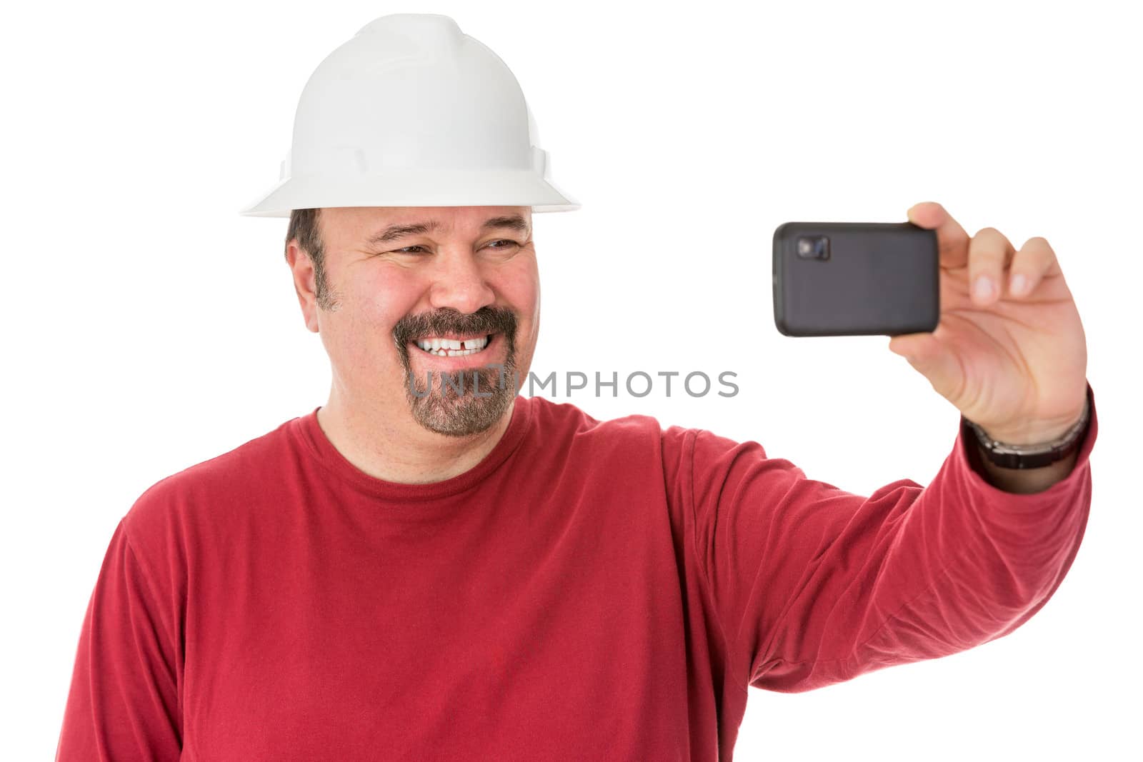 Workman with a neat goatee beard weaning a hardhat posing for a self-portrait giving a cheesy grin as he looks into the camera on his mobile phone, isolated on white