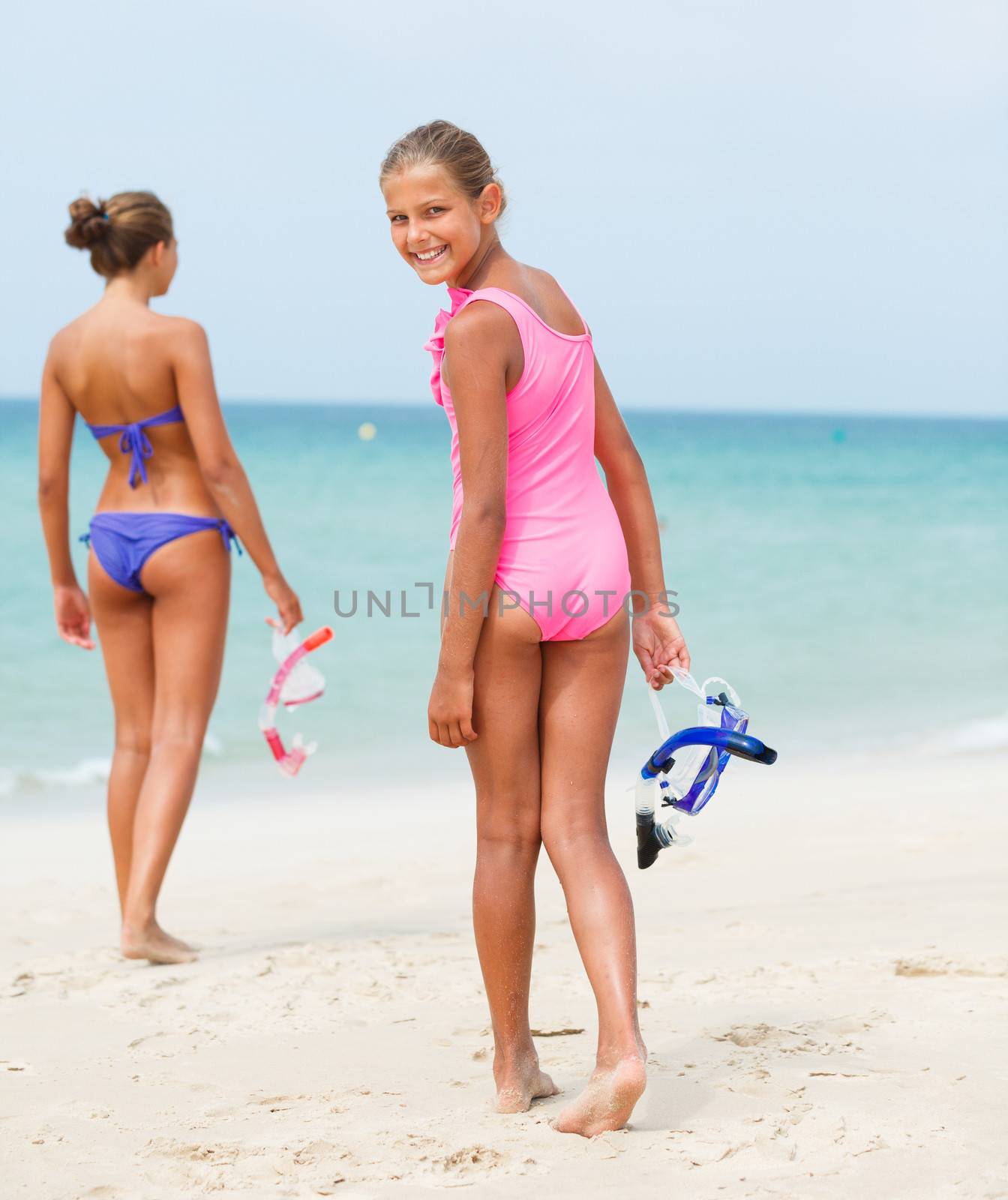 Back view of two happy girls on beach with colorful face masks and snorkels, sea in background. Vertical view.