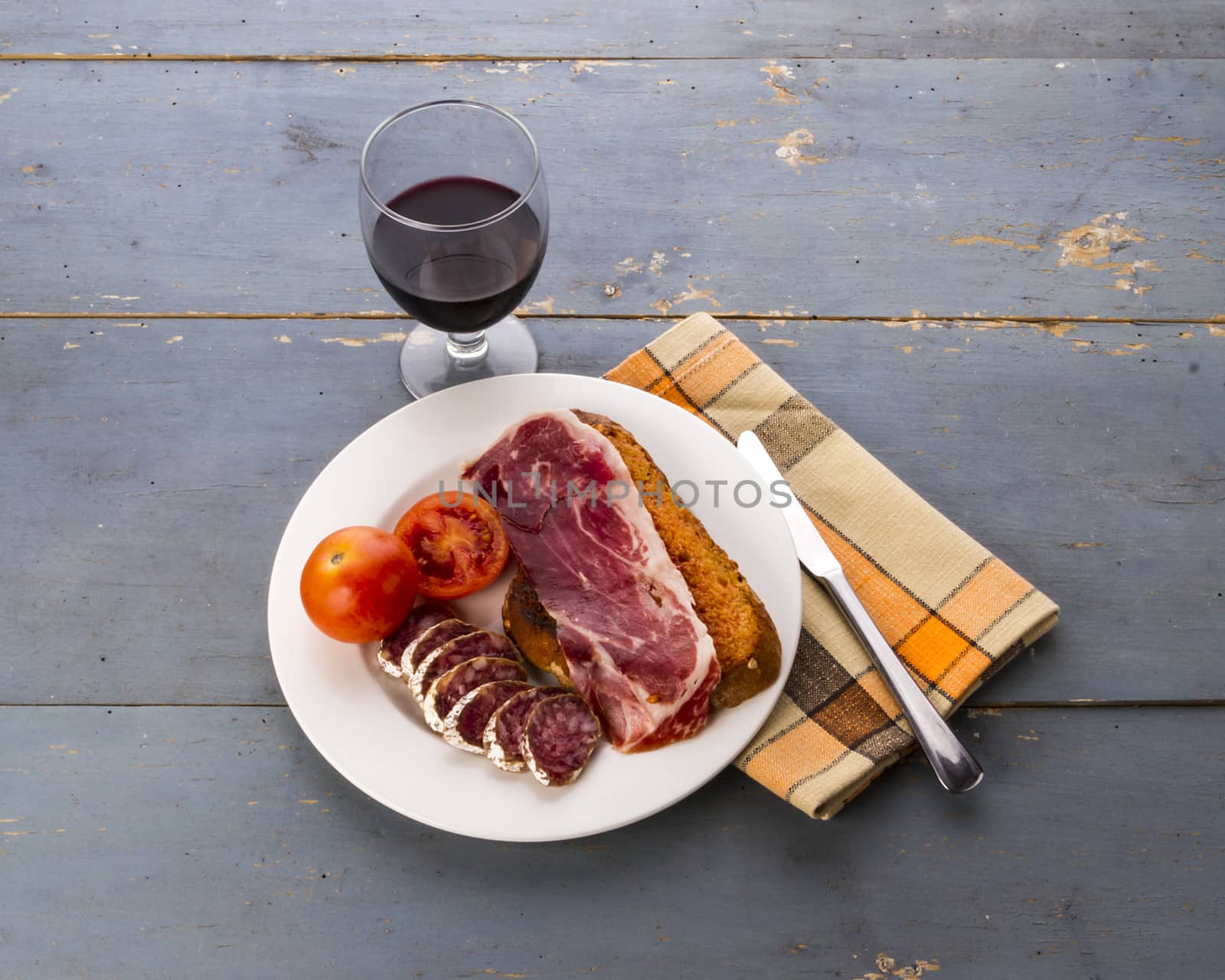 Still life with a catalan 'pa amb tomata' (bread or toast with tomato, salt and olive oil) with jam and cured meats, and a glass of red wine, on and old blue table