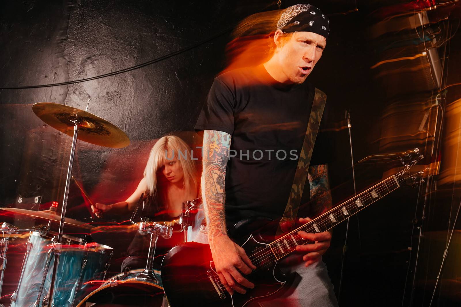 Band playing on a stage. Male guitarist and female drummer. Shot with strobes and slow shutter speed to create lighting atmosphere and blur effects. Slight motion blur on performers.