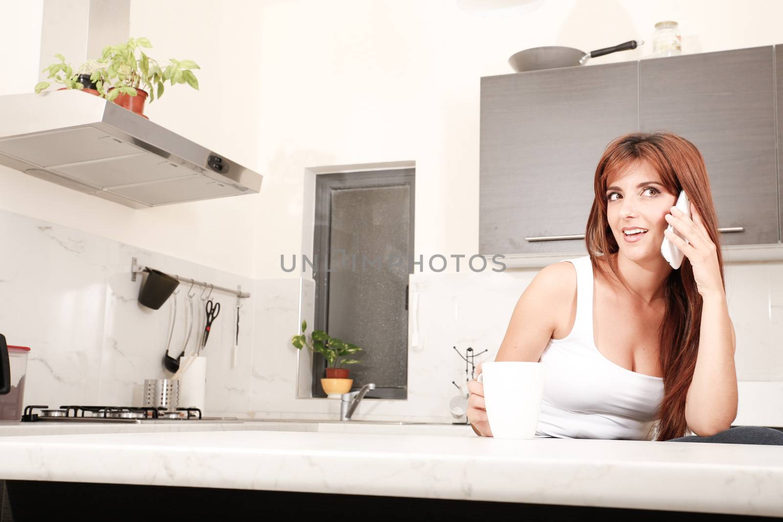 A young adult woman sitting in the kitchen while talking on a smartphone. 