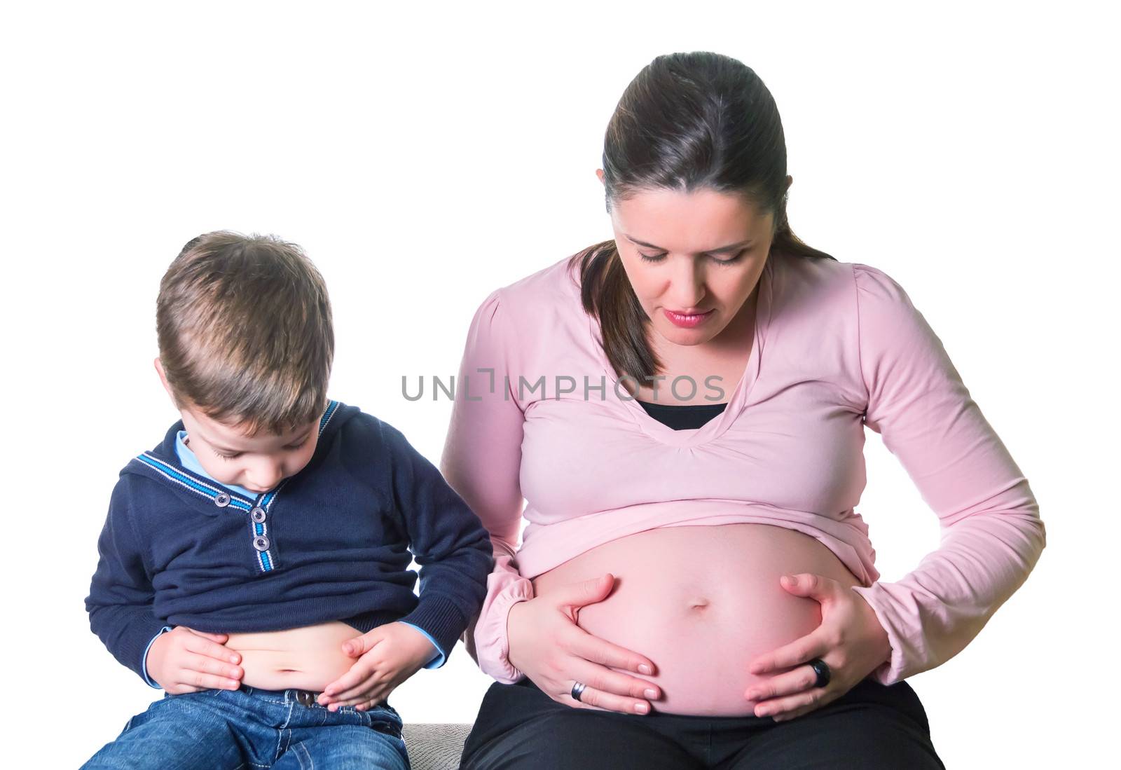 Pregnant mother and son laughing when comparing their bellies sitting on a sofa over white background