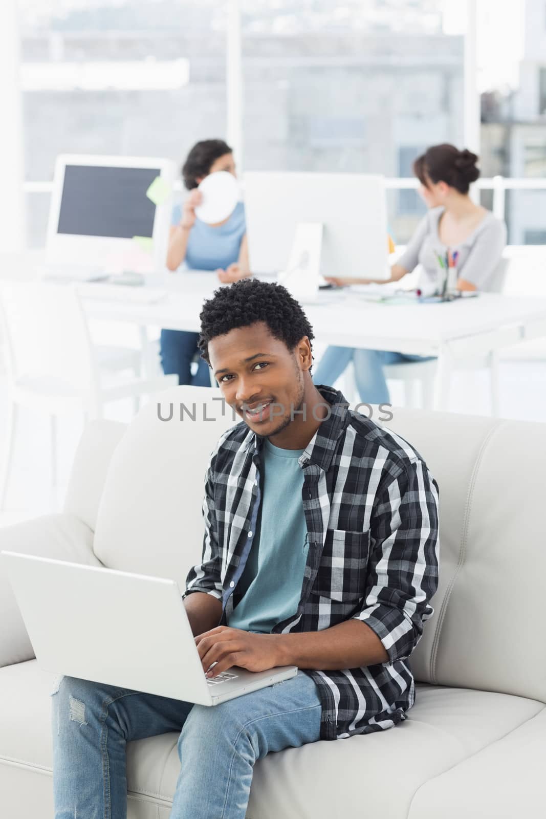 Young man using laptop with colleagues in background at a creative bright office