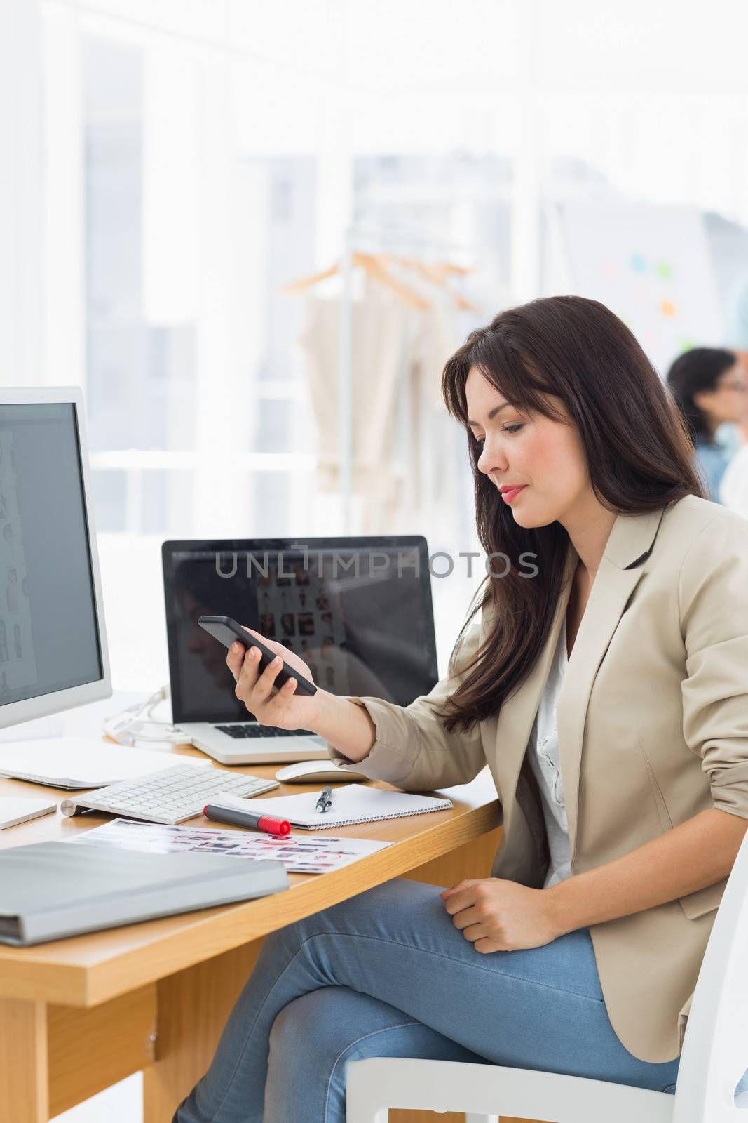 Casual young woman text messaging at desk with colleague in background at a bright office