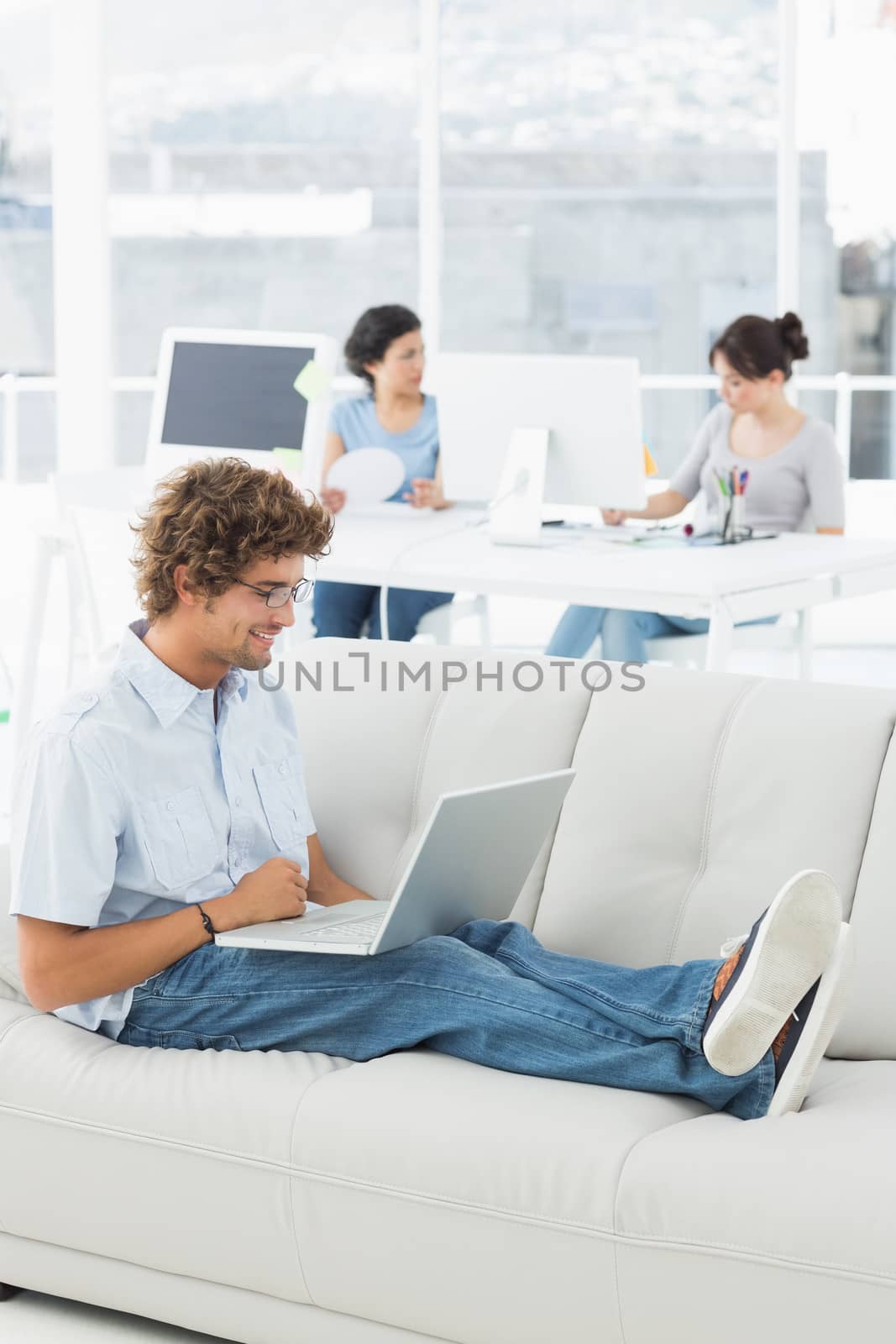 Young man using laptop with colleagues in background at a creative bright office