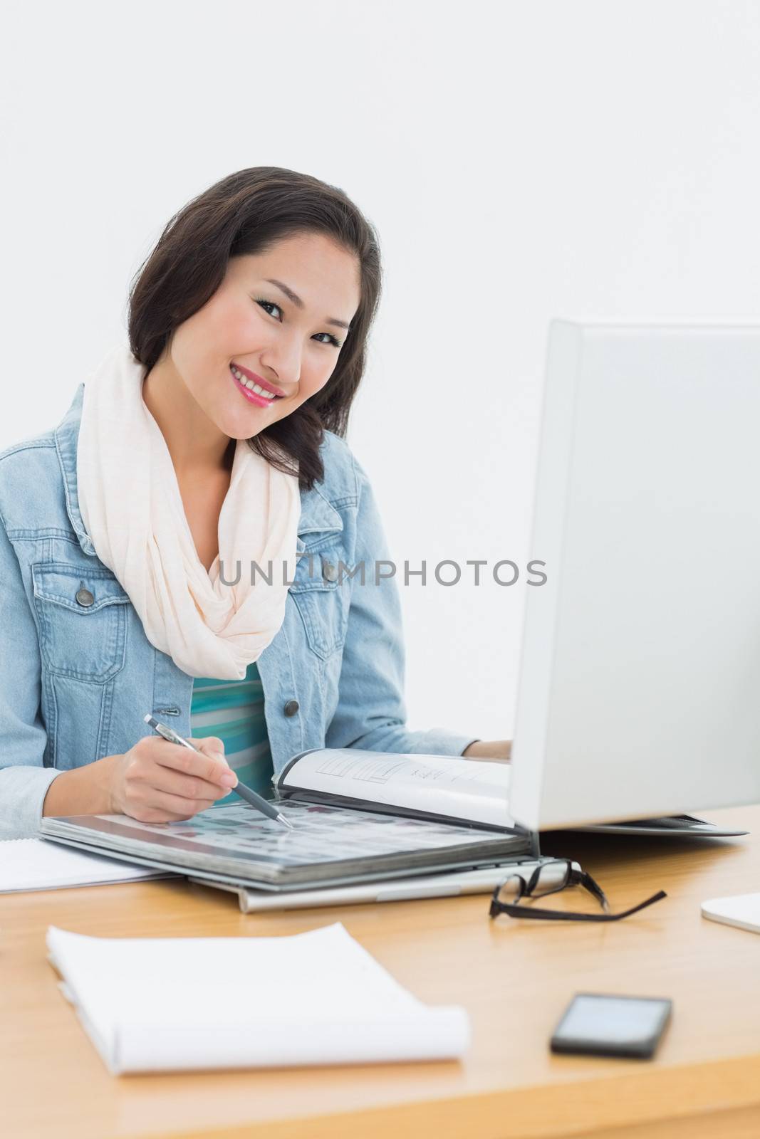 Portrait of a smiling casual young woman with catalog in front of computer in the office