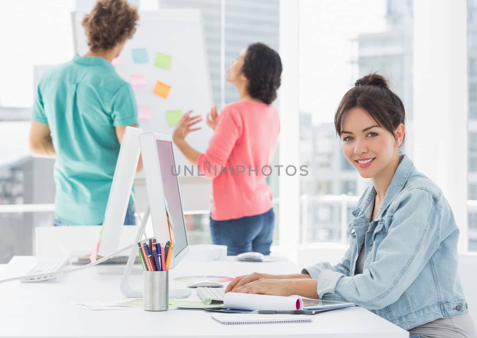 Side view portrait of a casual woman using computer with group of colleagues behind in a bright office