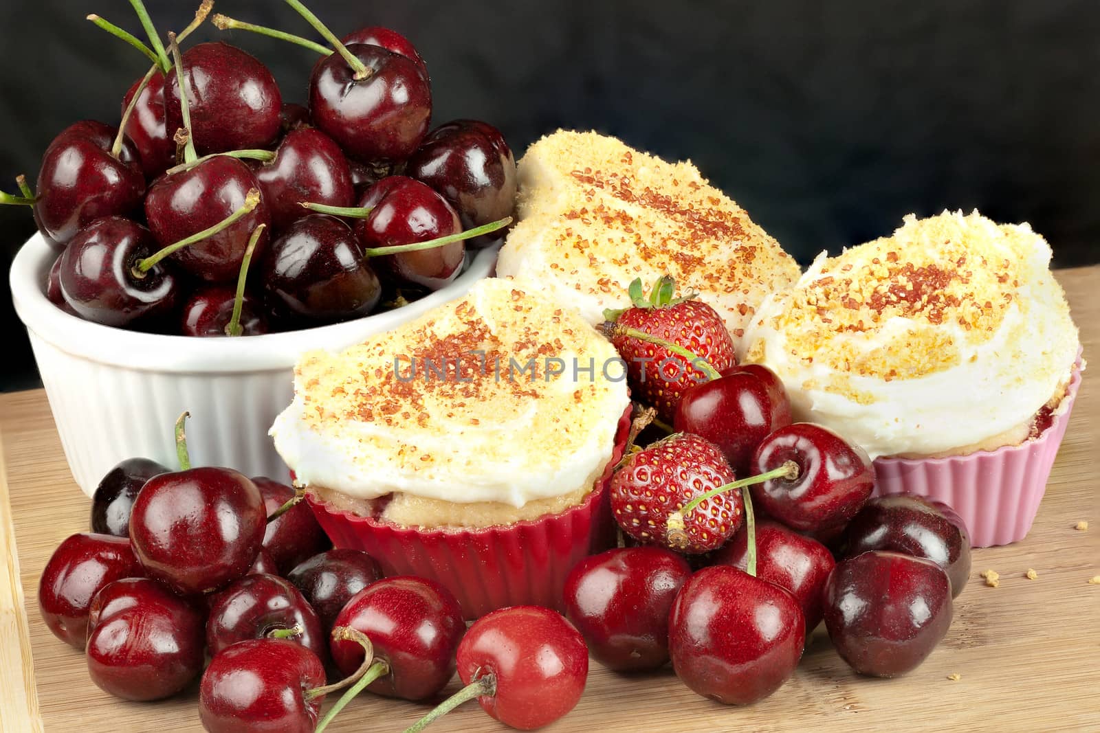 Close-up of cream cheese iced strawberry cherry muffins surrounded by fruit, topped with graham cracker crumbs.