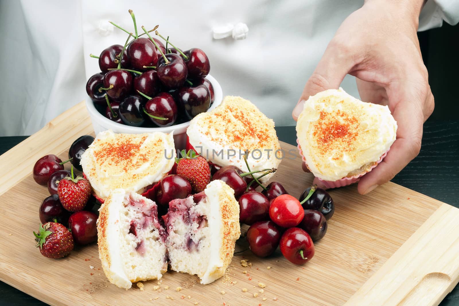 Close-up of a chef displaying a cream cheese iced strawberry cherry muffin surrounded by fruit and other muffins, topped with graham cracker crumbs.
