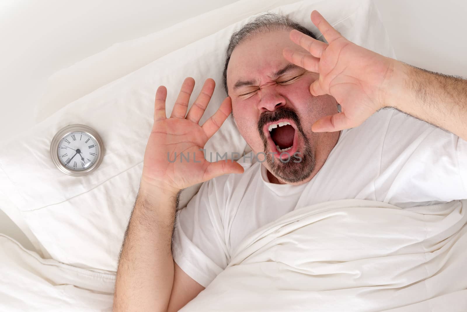 Middle-aged bearded man yawning loudly resting in bed with his alarm clock on the pillow alongside him as he tries to wake after a sleepless night