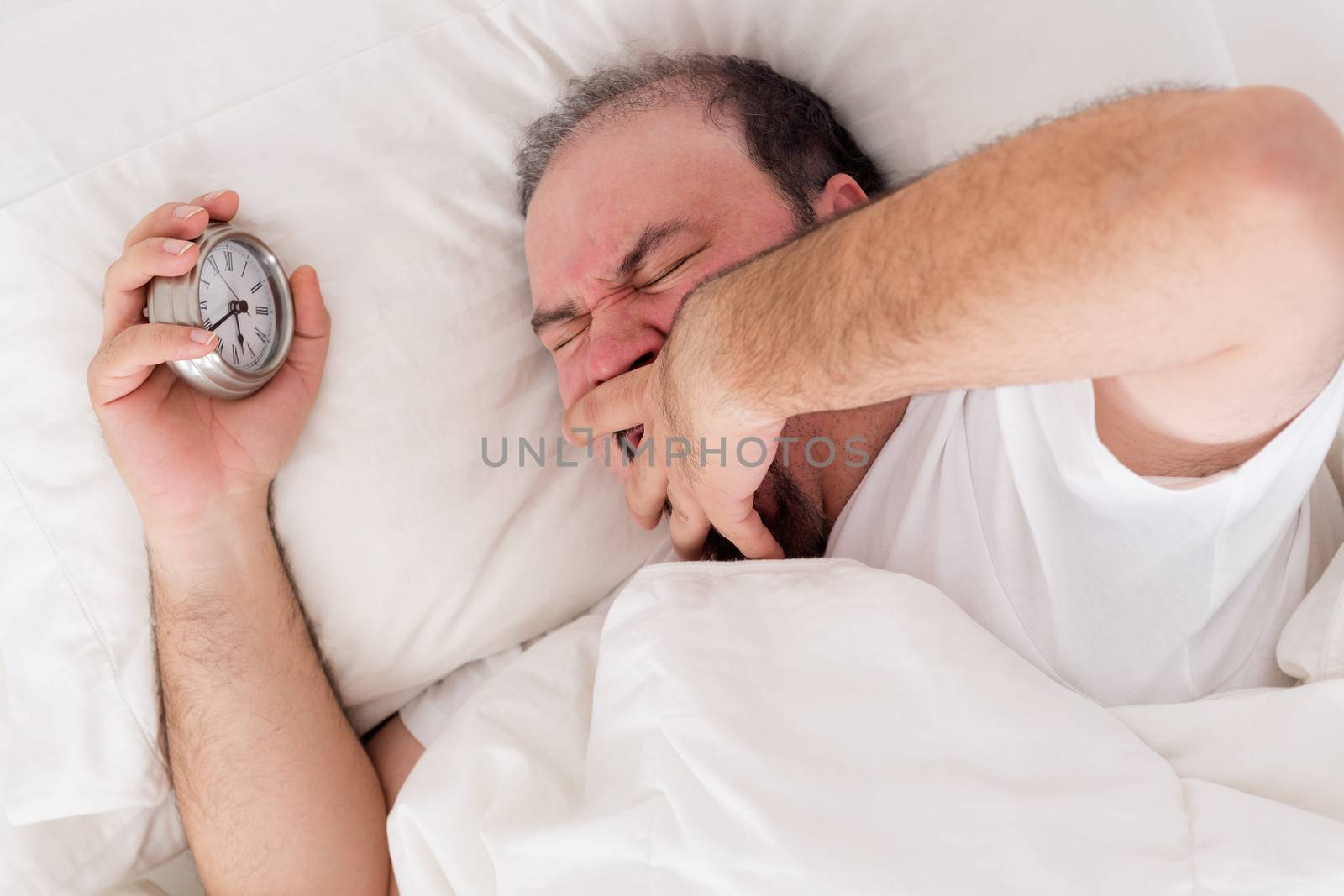 Man lying in bed yawning as he tries to wake up with his alarm clock clutched in his hand