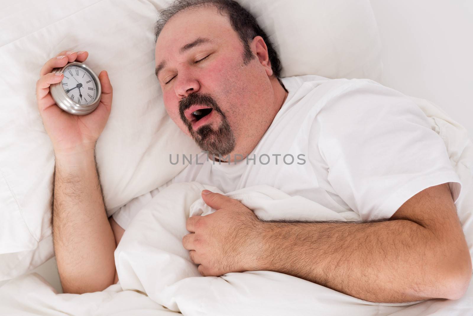 Lethargic tired man lying in bed yawning as he struggles to wake up unmotivated to start the new day and content to rather continue lying in bed as he holds his alarm clock in his hand